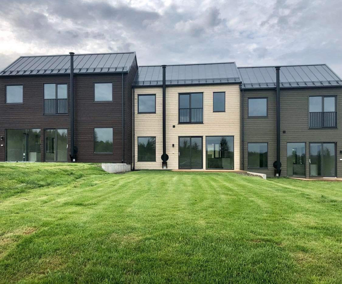 Trzy nowoczesne domy szeregowe z drewnianą elewacją w odcieniach brązu, beżu i zieleni, z metalowymi dachami i kominami, otoczone trawnikiem.