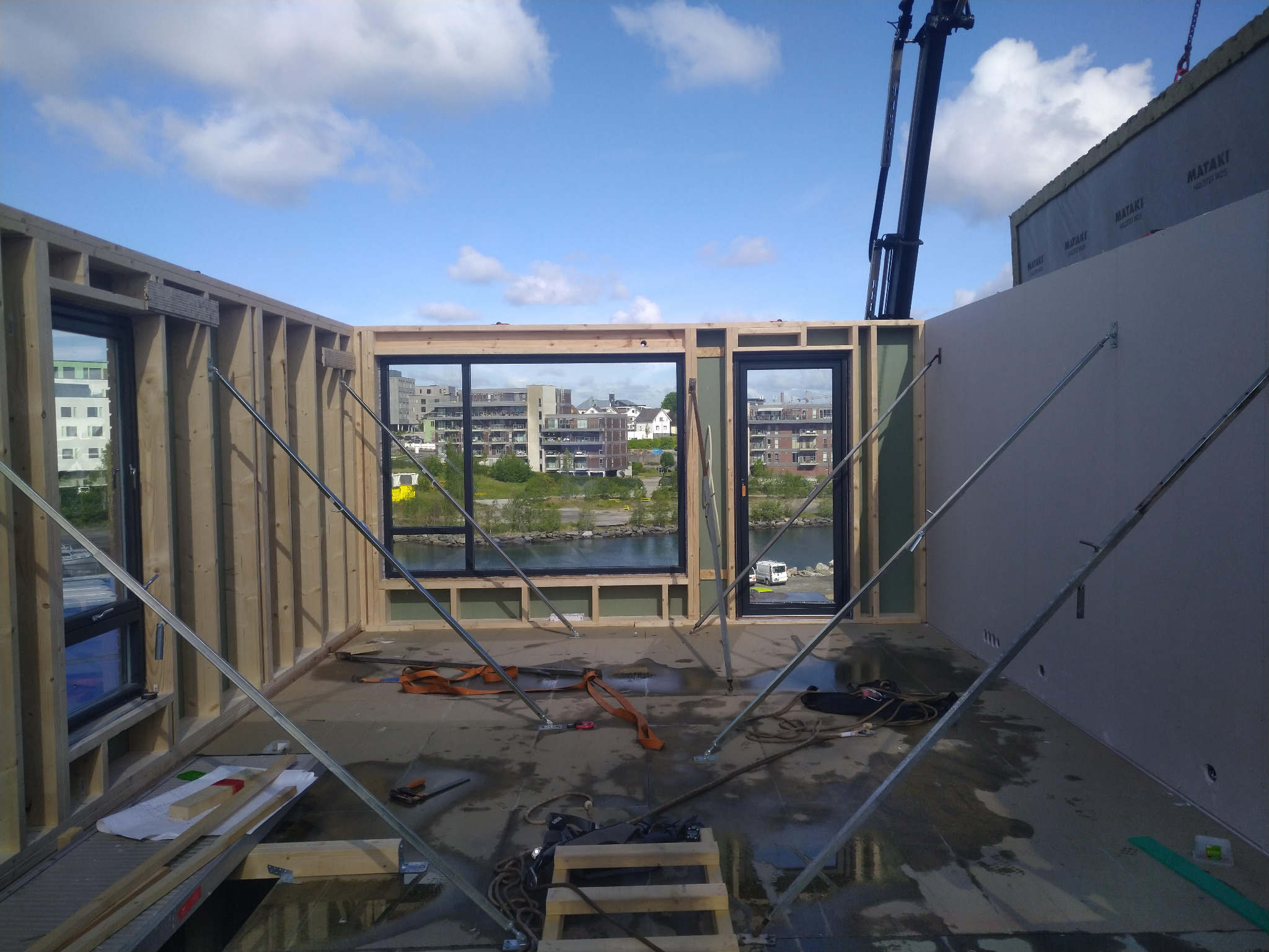 Wnętrze w trakcie budowy: drewniana konstrukcja szkieletowa z oknami, widok na miasto i rzekę, podpora stalowa, elementy mocujące, etap montażu ścian.