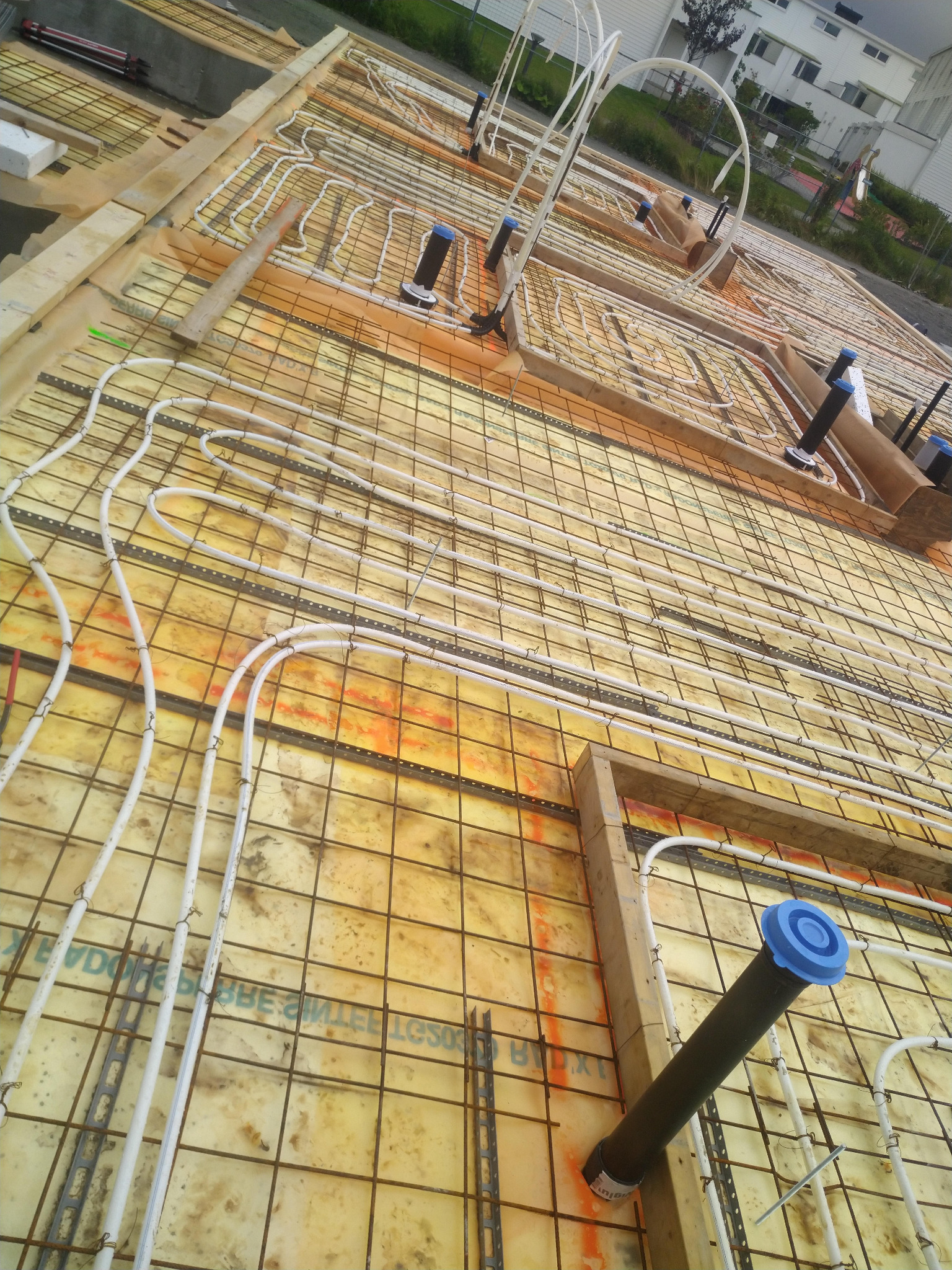 Instalacja ogrzewania podłogowego w trakcie budowy domu, widoczne rury grzewcze zamocowane do siatki zbrojeniowej na izolacji, z elementami pionowymi instalacji kanalizacyjnej.