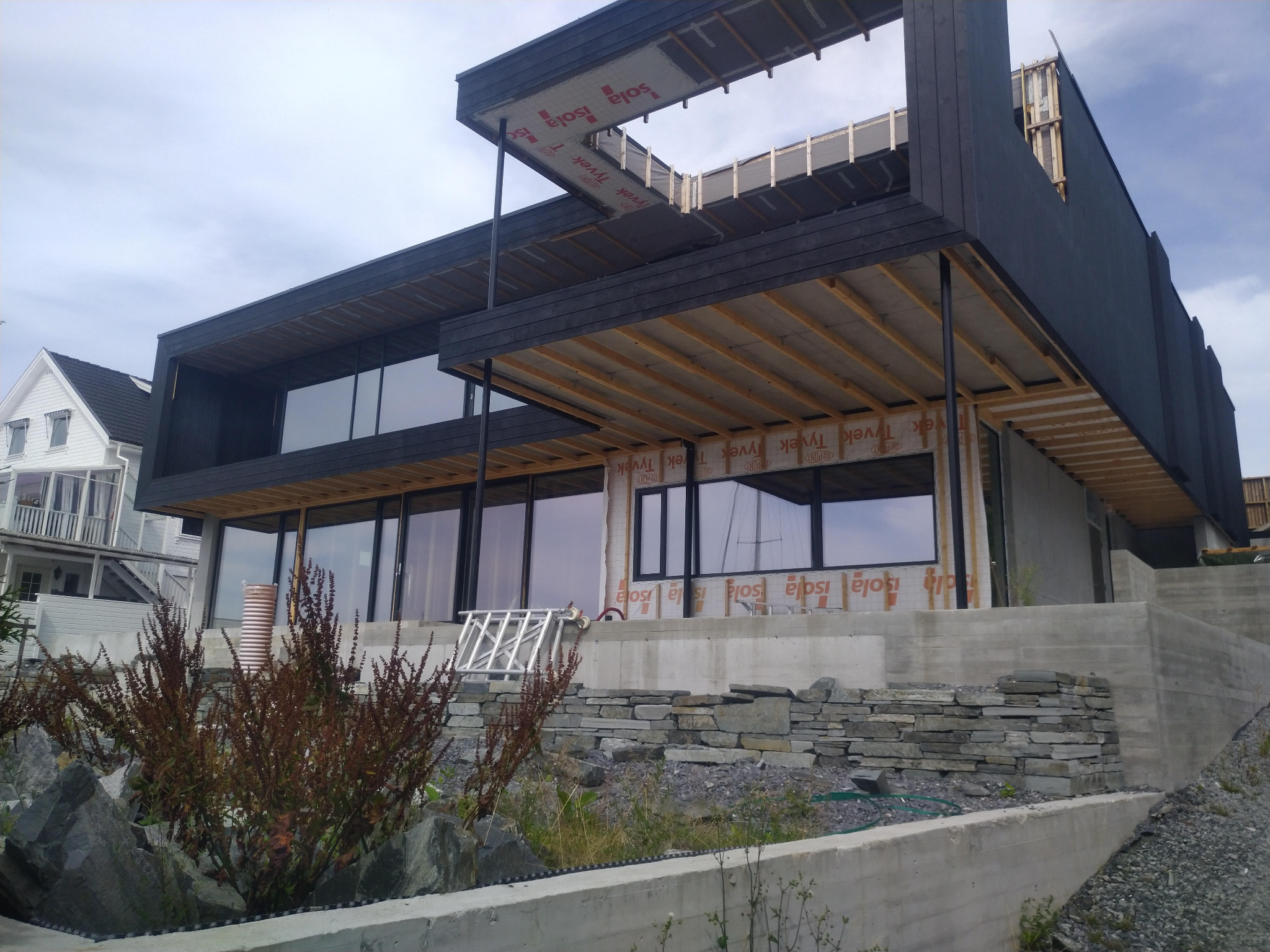 Nowoczesny dom szkieletowy w trakcie budowy z widoczną konstrukcją drewnianą, izolacją i dużymi oknami, na tle starszego domu.