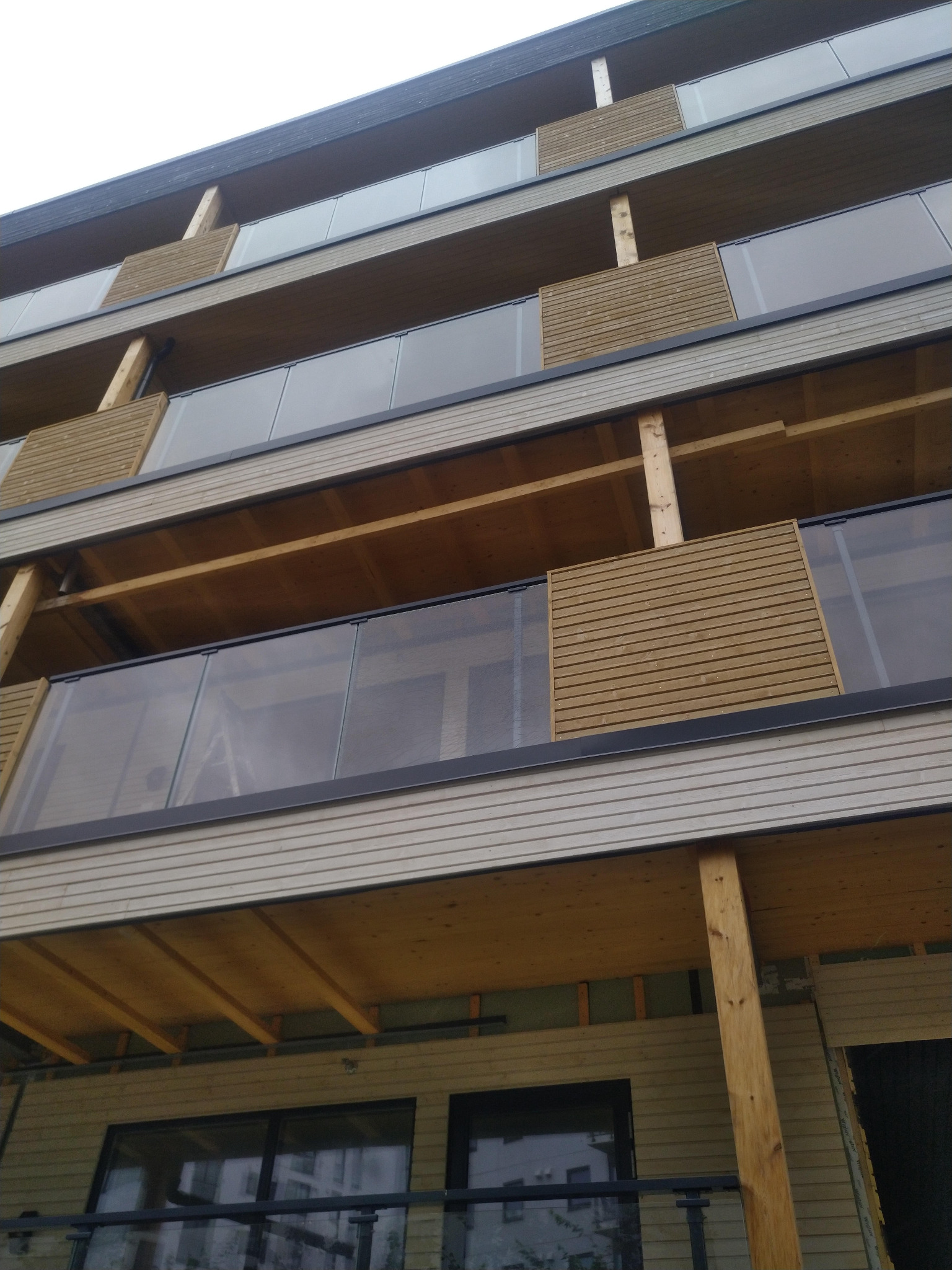 Wielopiętrowy budynek z drewnianą elewacją, balustradami z przezroczystego szkła i drewnianymi panelami na balkonach, widziany z dołu pod kątem.