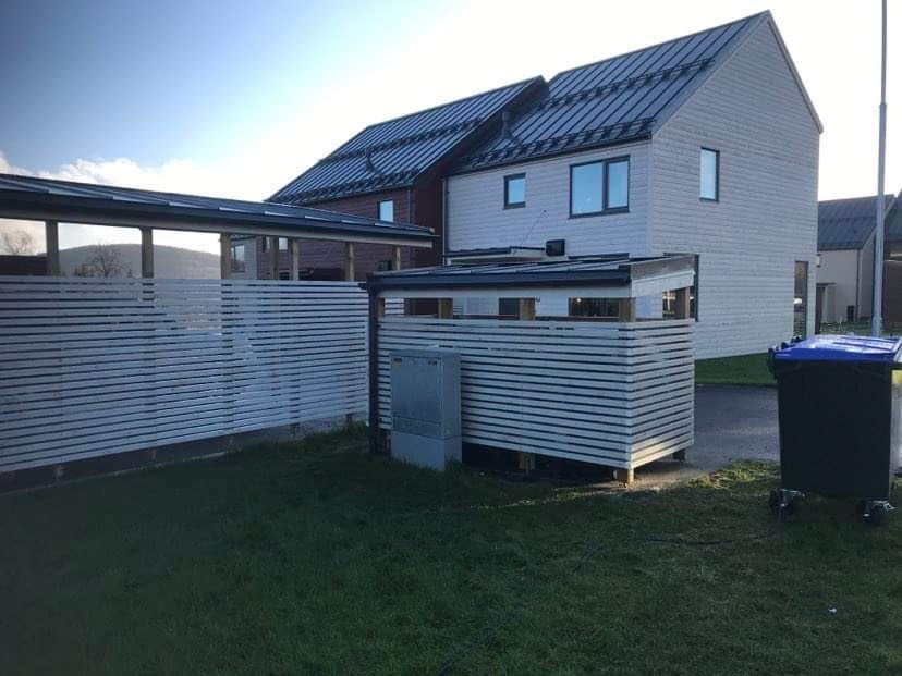 Nowoczesny dom z białymi panelami elewacyjnymi i ciemnym dachem, z widoczną osłoną na śmietnik wykonaną z poziomych desek i niebieskim pojemnikiem na śmieci na trawniku.