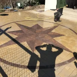 Umyta kostka brukowa z wzorem gwiazdy w kolorach beżu i brązu, z cieniem osoby trzymającej aparat i osoby spłukującej powierzchnię wodą z węża.