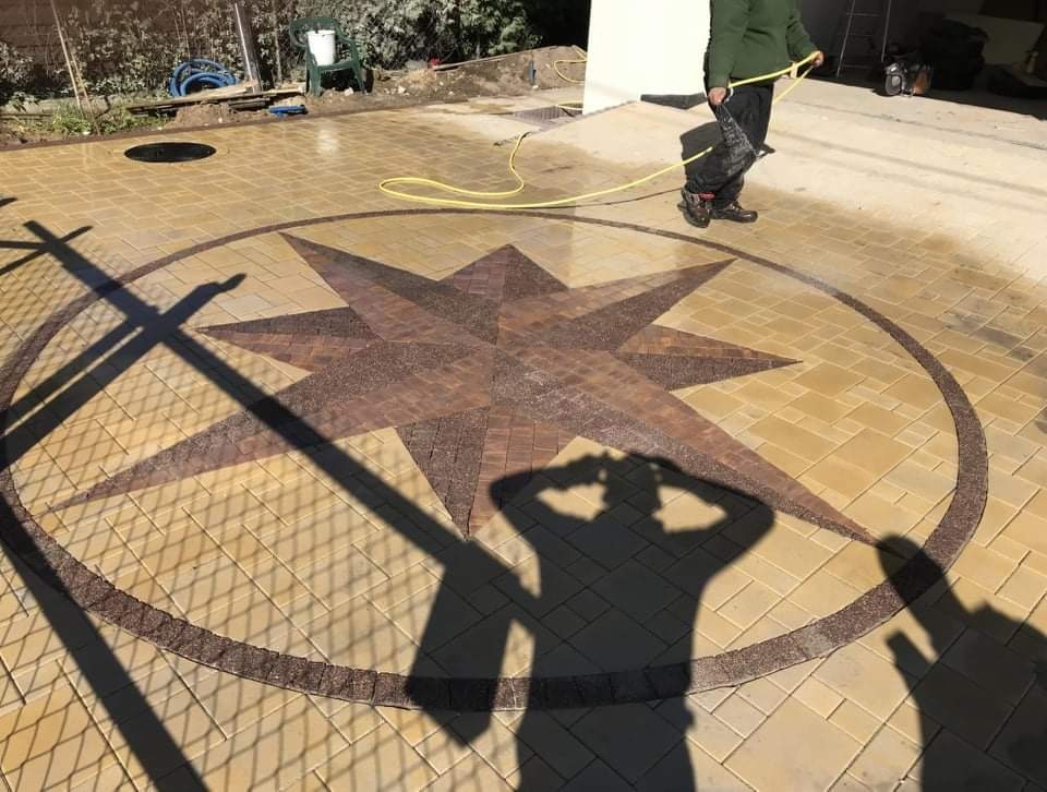 Umyta kostka brukowa z wzorem gwiazdy w kolorach beżu i brązu, z cieniem osoby trzymającej aparat i osoby spłukującej powierzchnię wodą z węża.