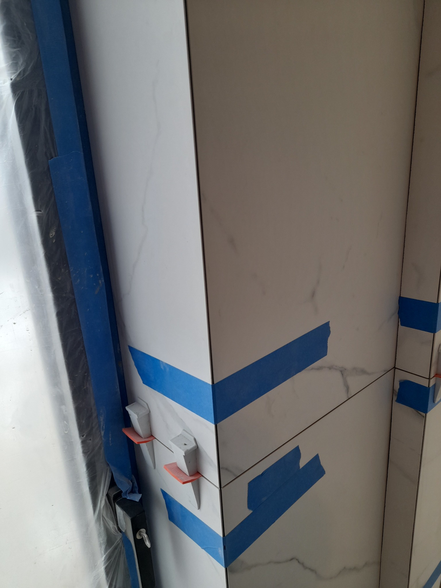 Ściana wyłożona białymi płytkami imitującymi marmur, zabezpieczona niebieską taśmą malarską i poziomowana za pomocą klinów dystansowych podczas prac wykończeniowych.