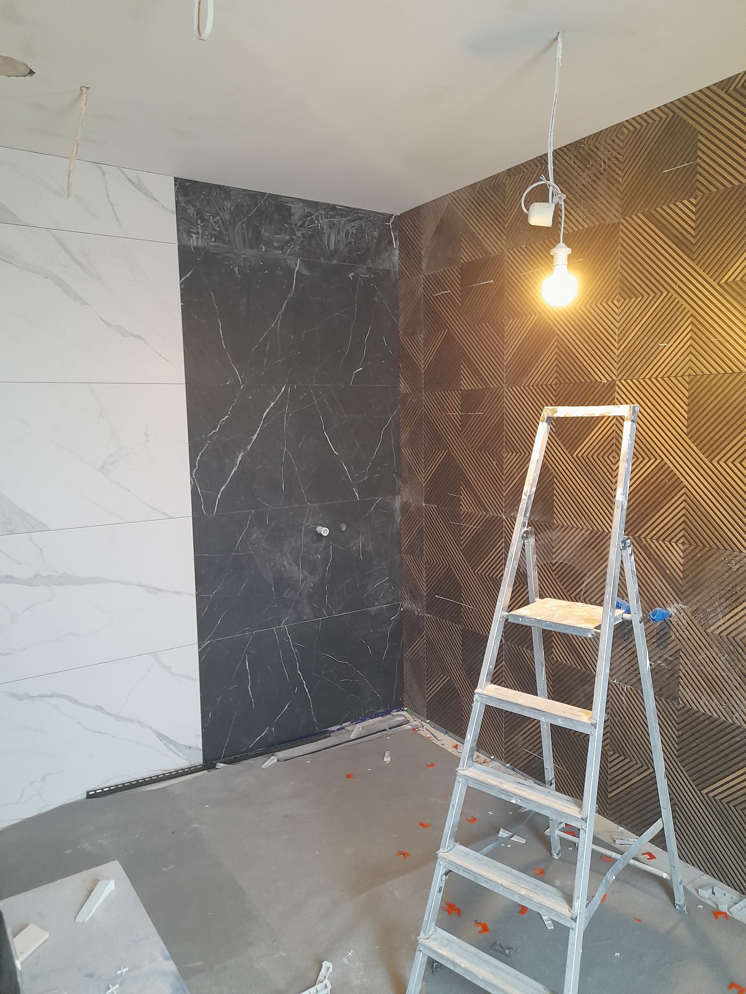 Remont łazienki w trakcie: ułożone płytki imitujące marmur w kolorze białym i czarnym, dekoracyjna ściana z geometrycznym wzorem, drabina aluminiowa i wisząca żarówka.