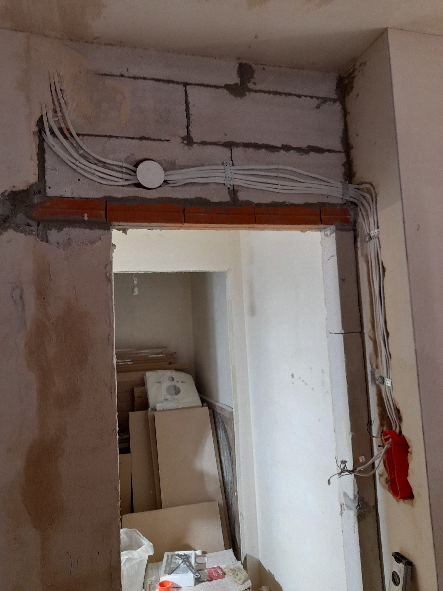 Instalacja elektryczna w trakcie remontu: białe przewody ułożone wzdłuż surowego muru nad otworem drzwiowym, widoczna puszka instalacyjna i narzędzia w tle.