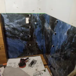 Fragment łazienki w trakcie układania płytek imitujących marmur w odcieniach granatu i błękitu, widoczne krzyżyki dystansowe, szlifierka kątowa, paca i kartony z logo Grohe.