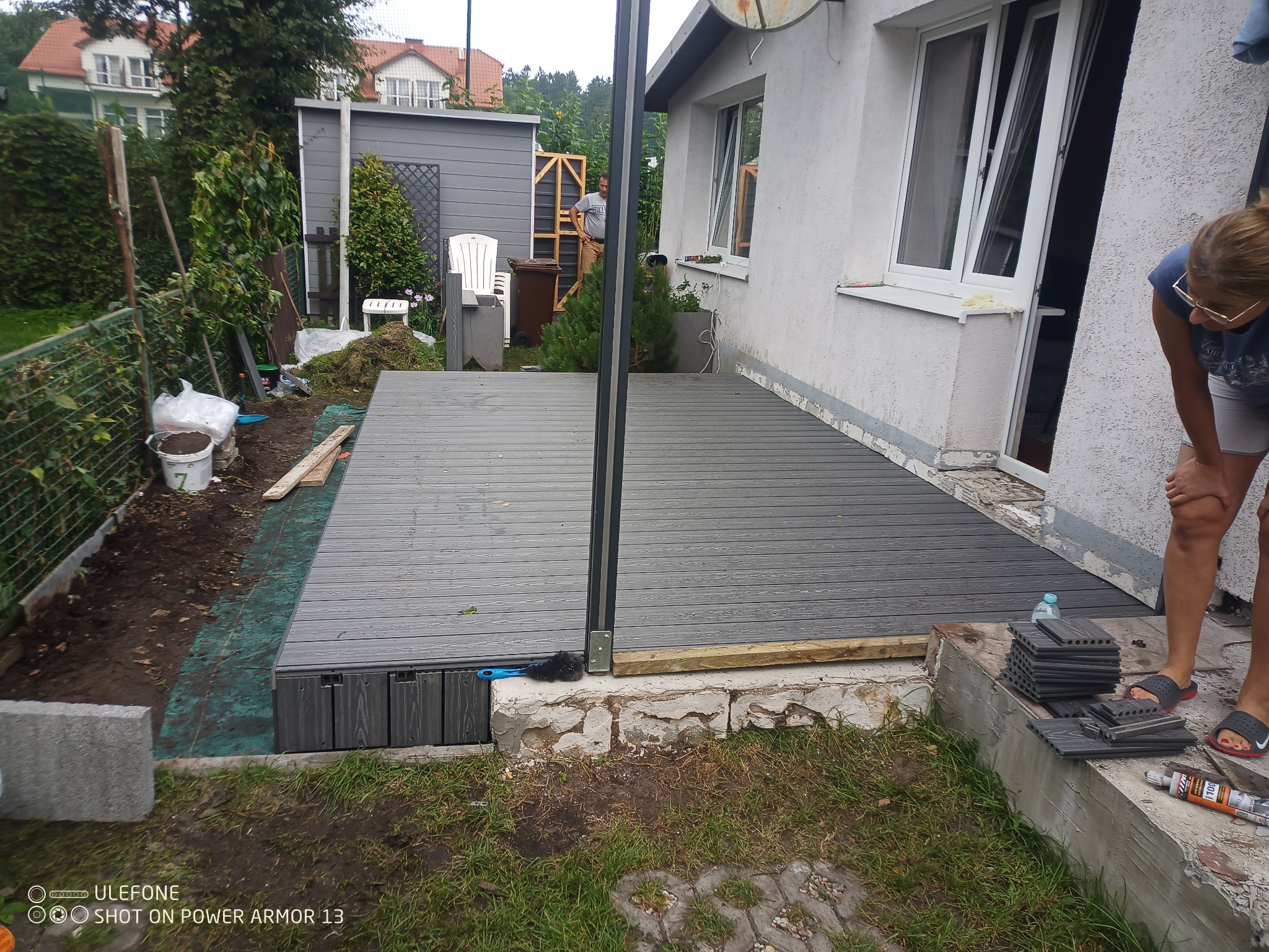 Montaż kompozytowego tarasu w ogrodzie: widoczne deski tarasowe, elementy konstrukcyjne, narzędzia i osoba przy pracy, z zielenią w tle.