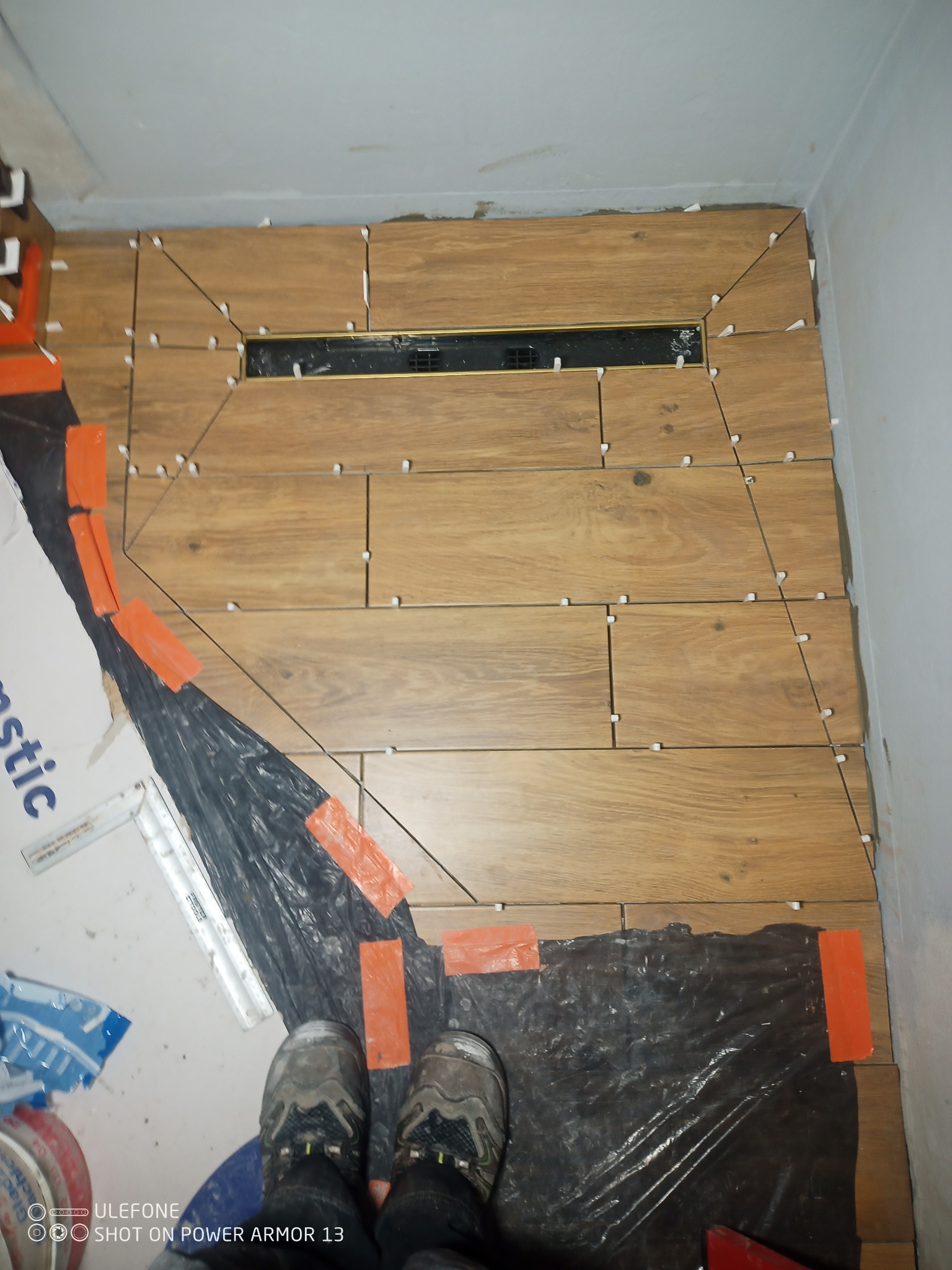 Układanie płytek imitujących drewno w łazience, widoczny odpływ liniowy i krzyżyki dystansowe, fragment folii ochronnej i robocze buty wykonawcy.