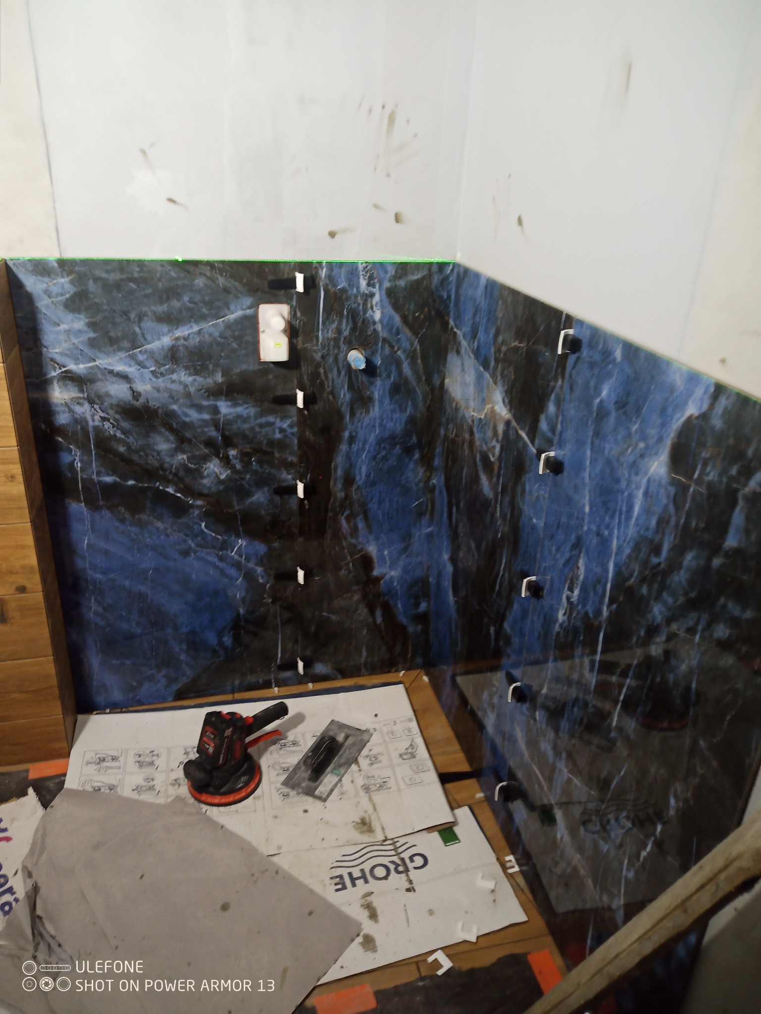 Fragment łazienki w trakcie układania płytek imitujących marmur w odcieniach granatu i błękitu, widoczne krzyżyki dystansowe, szlifierka kątowa, paca i kartony z logo Grohe.