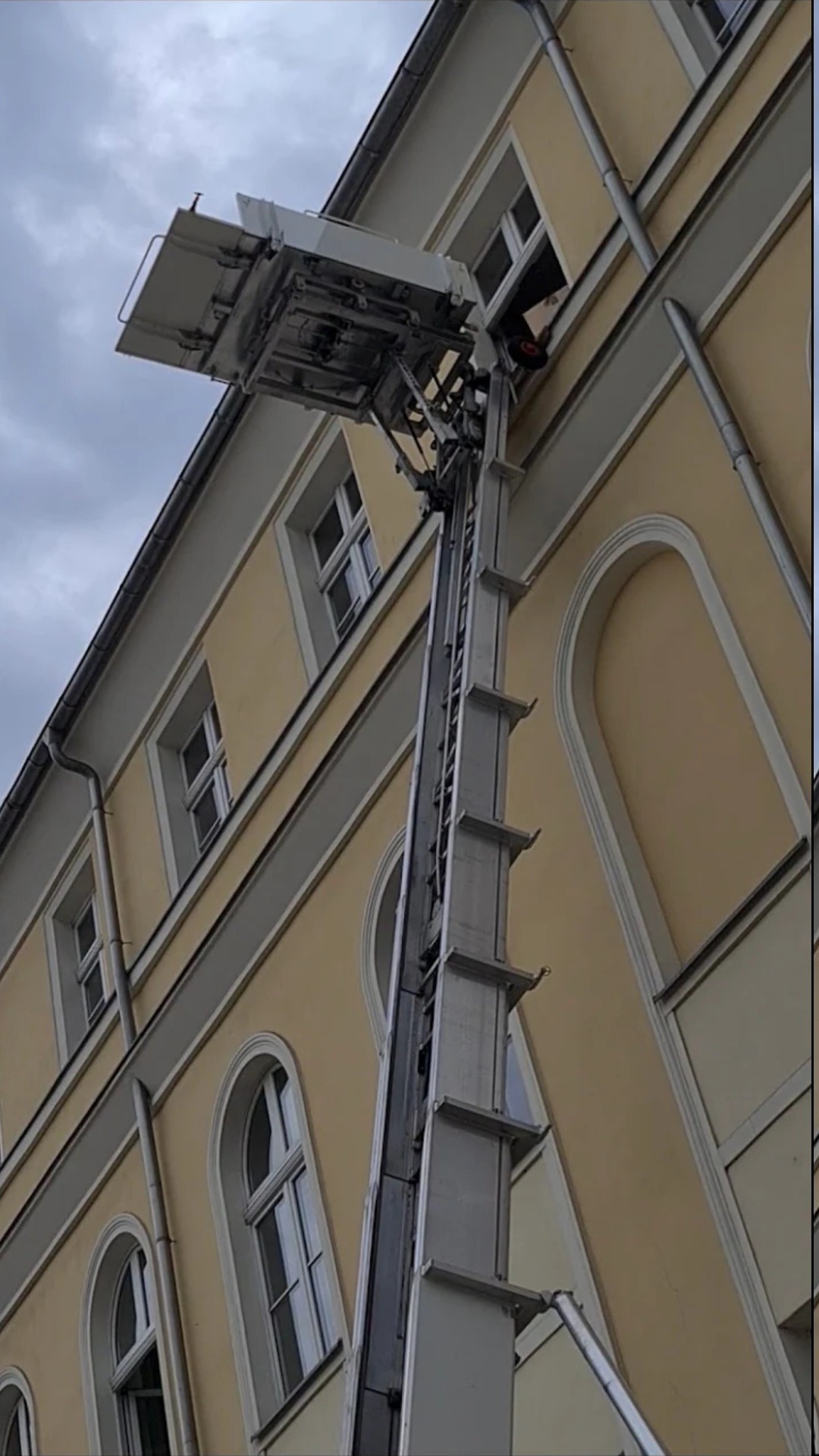 Zewnętrzny podnośnik meblowy sięga do okna budynku o żółtej elewacji, widoczny fragment sylwetki pracownika w oknie.