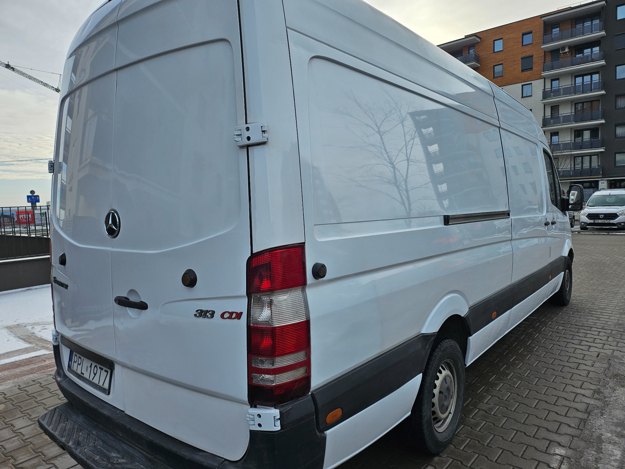 Biały samochód dostawczy Mercedes Sprinter 313 CDI z tablicą rejestracyjną PPL 19T7 zaparkowany na brukowanej ulicy w Żelazkowie, w tle widoczny budynek mieszkalny z balkonami.