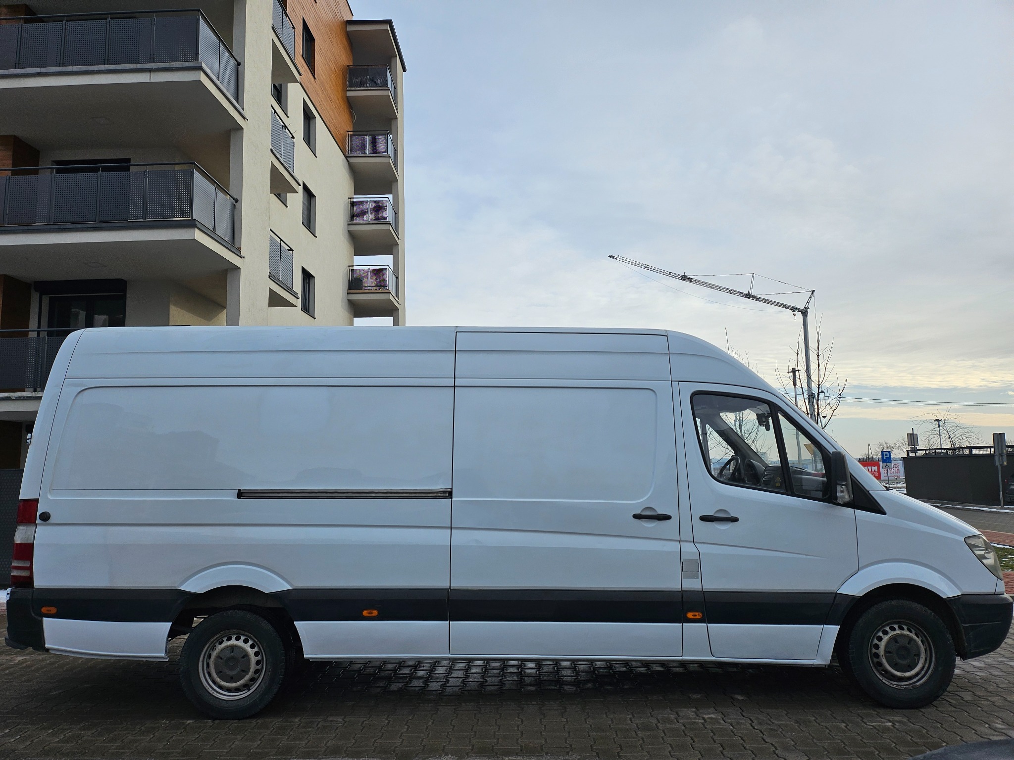 Biały furgon dostawczy zaparkowany na brukowanej ulicy przed nowoczesnym budynkiem mieszkalnym z balkonami, z żurawiem budowlanym widocznym w tle.