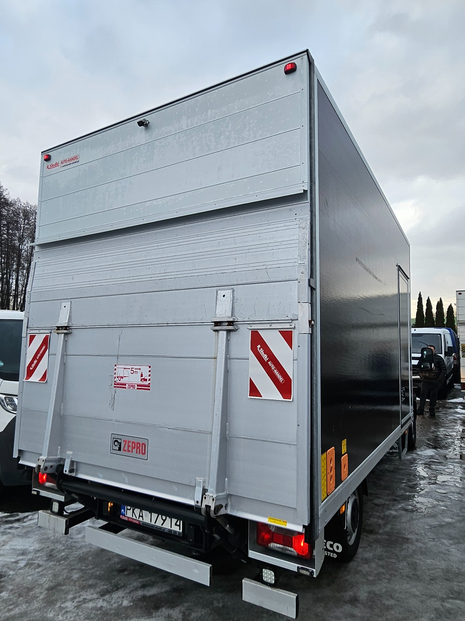 Zamknięta skrzynia ładunkowa samochodu dostawczego z windą załadowczą Zepro, widoczne tablice ostrzegawcze i rejestracja PKA 17914, na mokrym placu manewrowym.