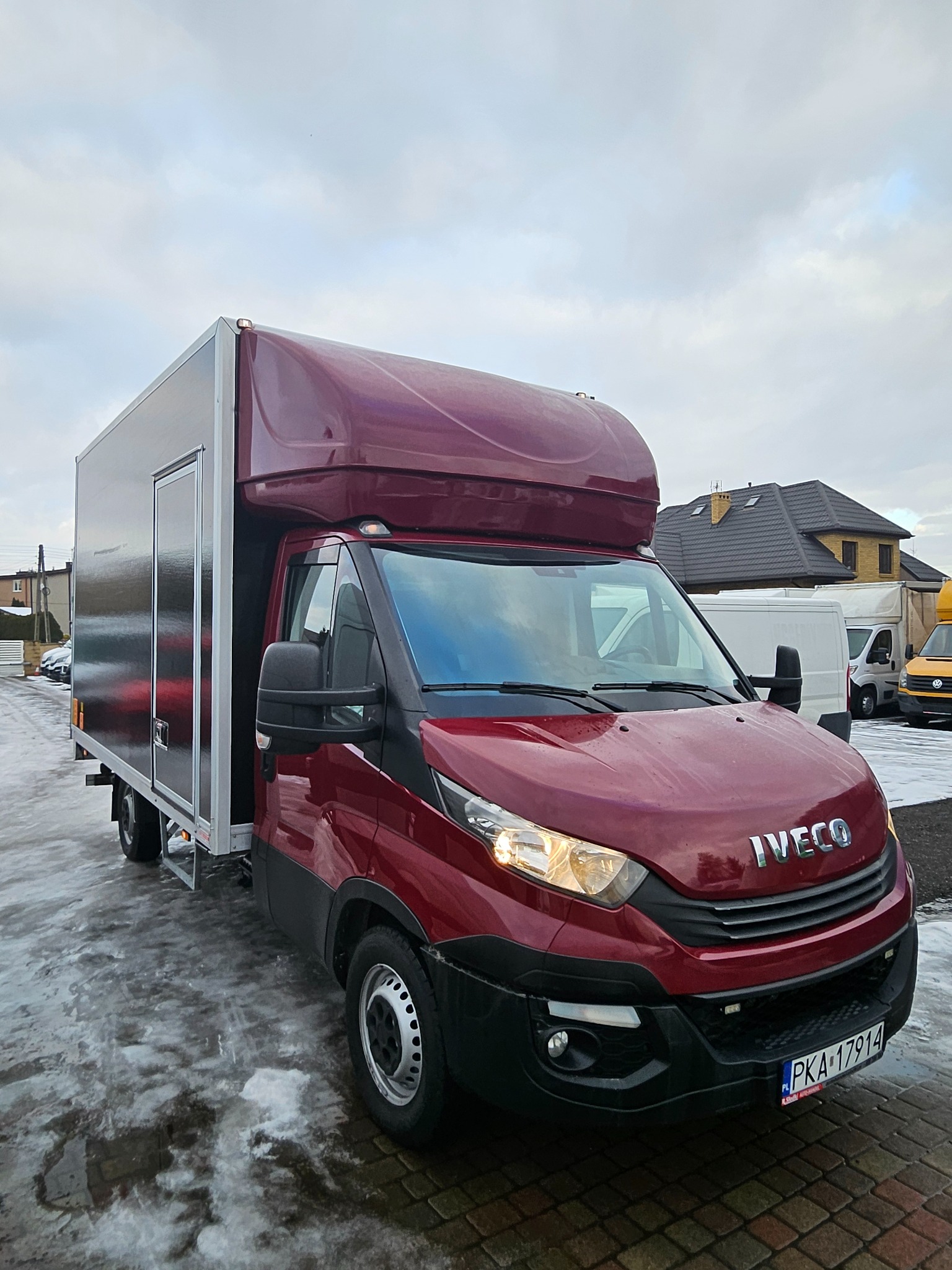 Czerwony samochód dostawczy Iveco z zabudową kontenerową i aluminiowymi drzwiami, zaparkowany na mokrym, częściowo ośnieżonym placu.