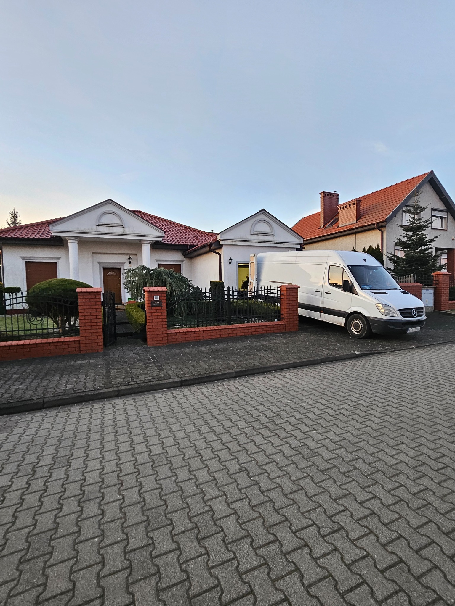 Biały samochód dostawczy zaparkowany przed domem z czerwonym dachem i ceglanym ogrodzeniem. Widoczna brama wjazdowa i część budynku sąsiada.