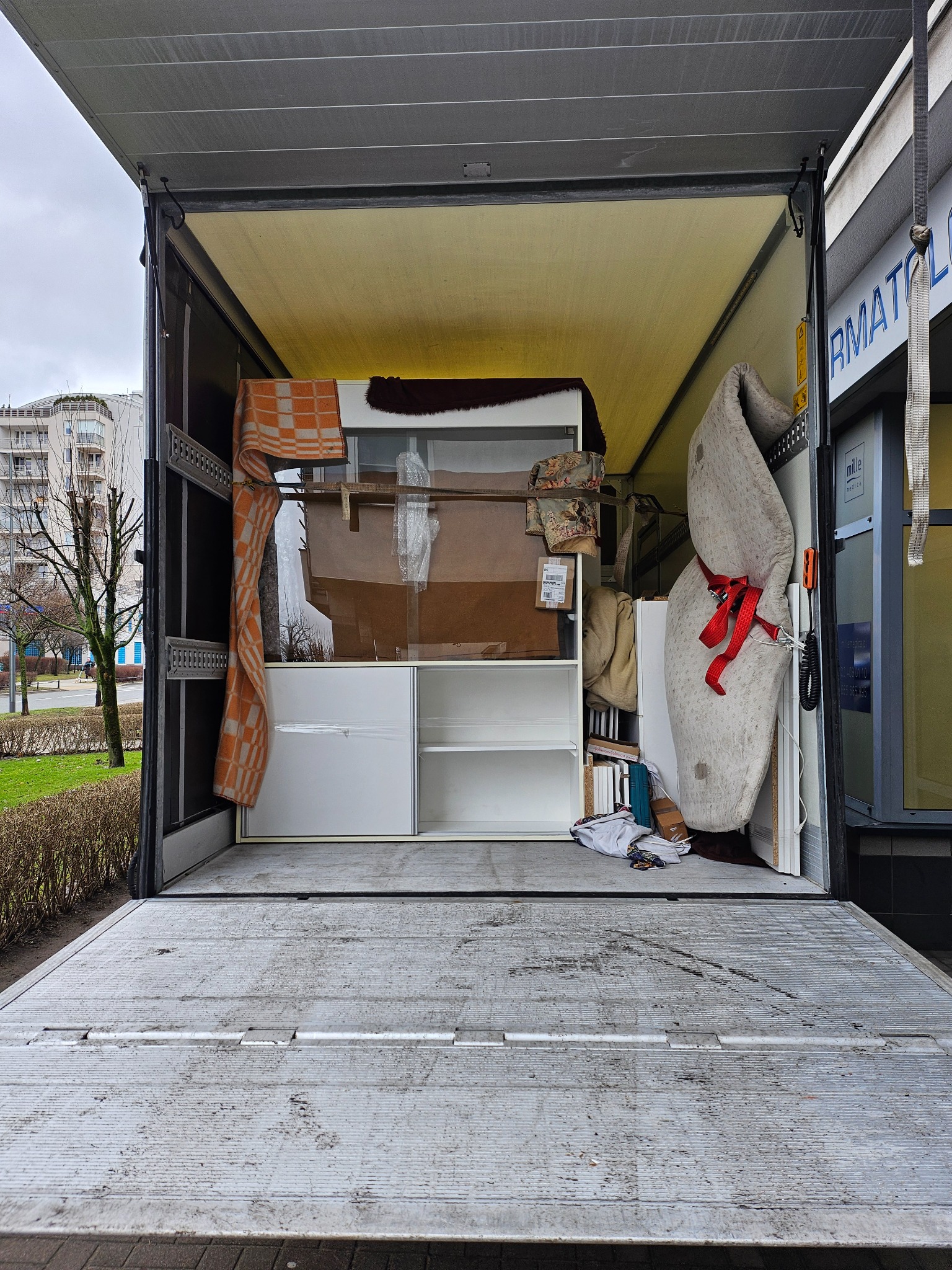 Wnętrze otwartej ciężarówki wypełnione meblami i przedmiotami zabezpieczonymi do transportu, widoczny fragment ulicy z budynkami w tle.