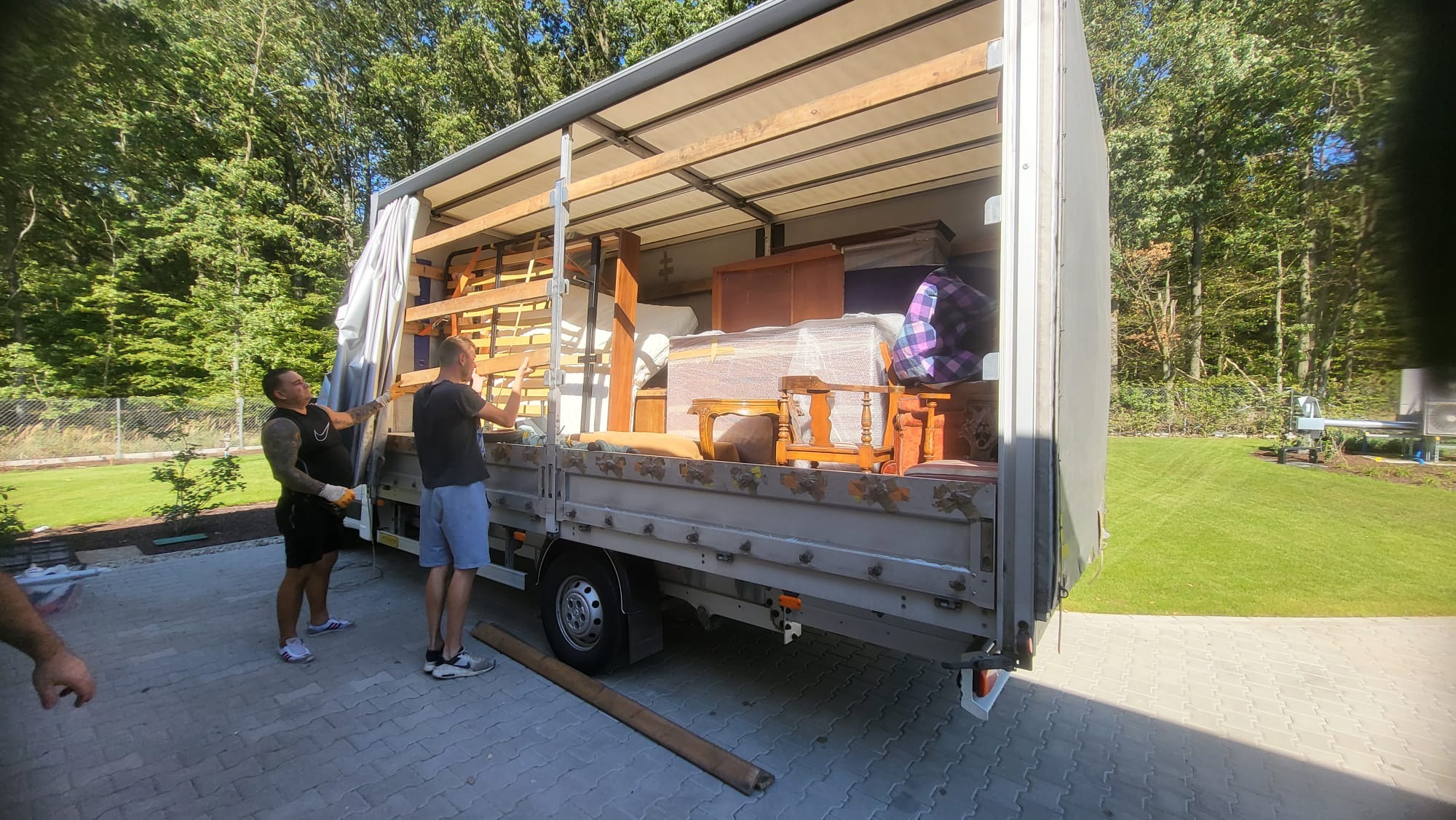 Dwóch mężczyzn ładuje meble, w tym drewniane krzesła i komodę, do ciężarówki z otwartą przestrzenią ładunkową, na tle zielonego trawnika i drzew.