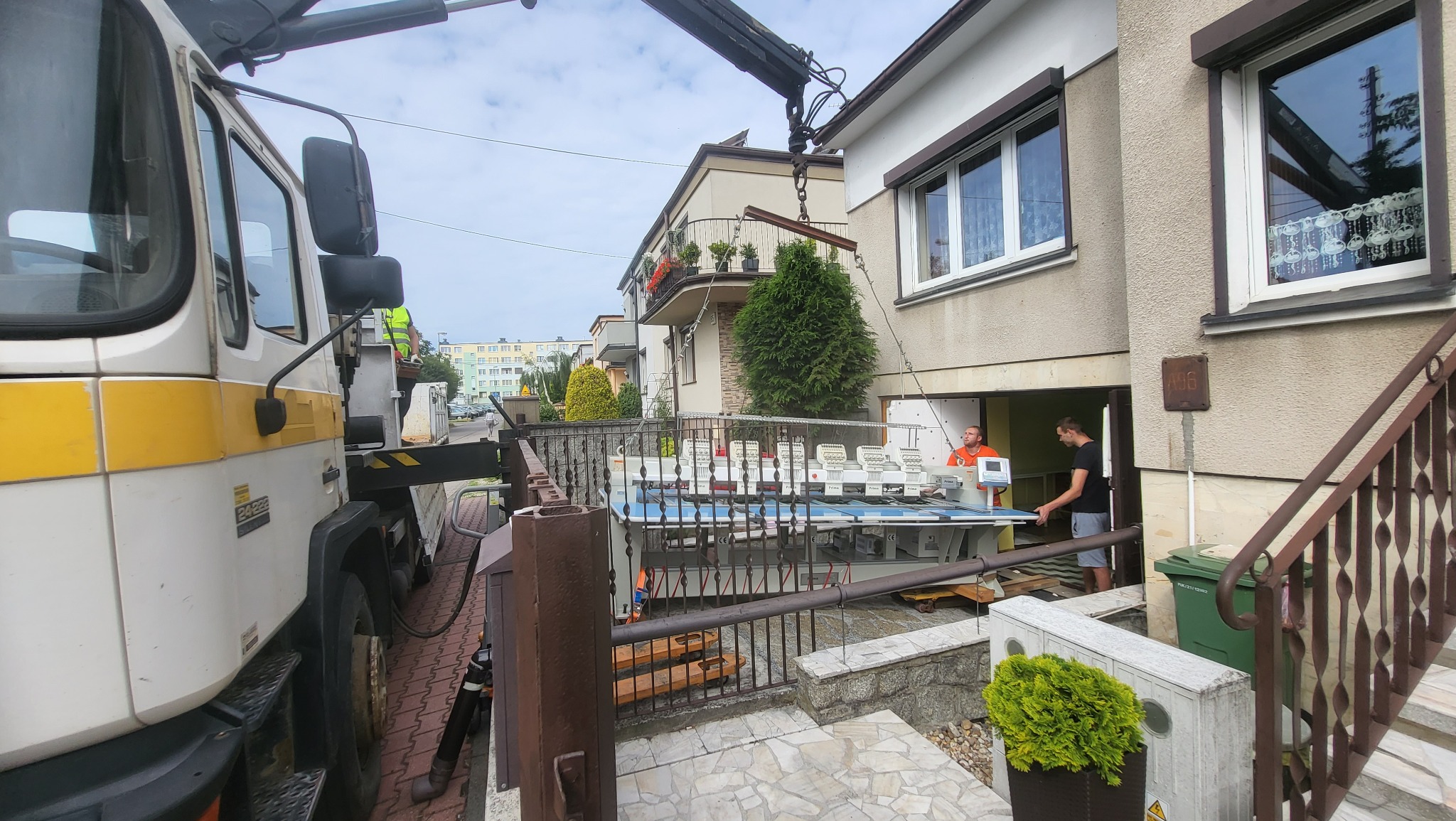 Dźwig transportuje dużą maszynę przemysłową do wnętrza budynku, pracownicy nadzorują proces, widoczny fragment ciężarówki z żółtymi pasami.