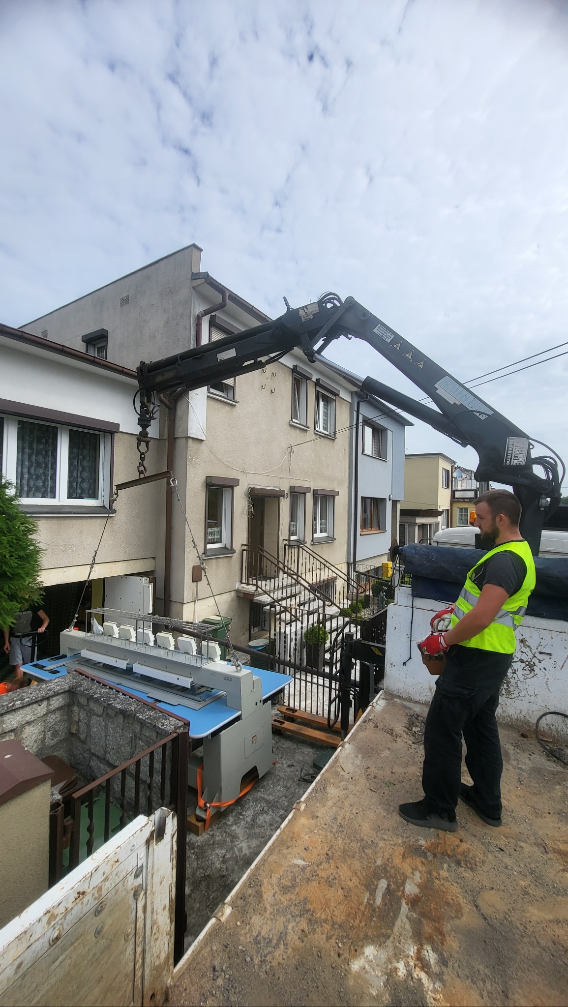 Przenoszenie maszyny przemysłowej dźwigiem na tle budynku mieszkalnego, pracownik w kamizelce odblaskowej steruje operacją.
