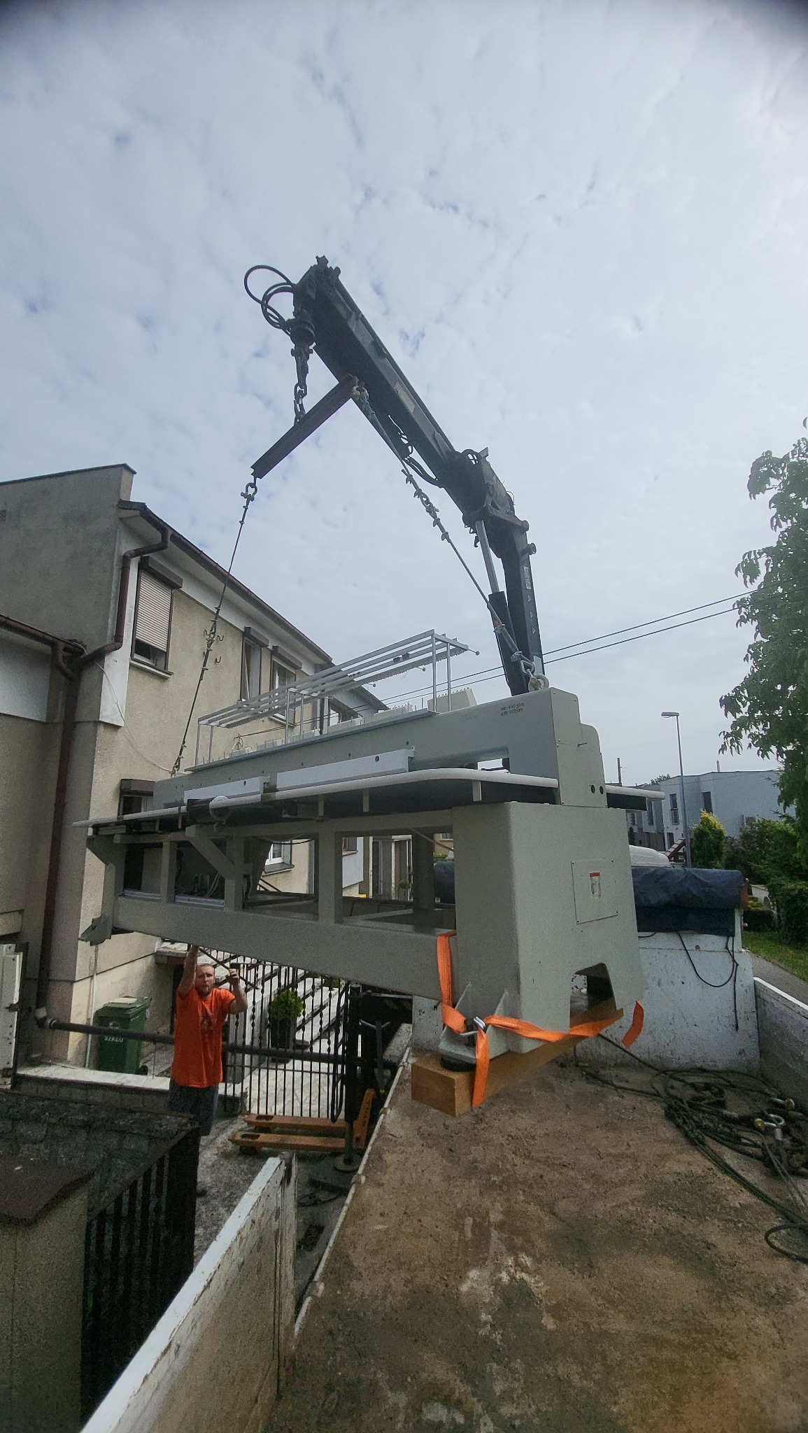 Dźwig podnosi dużą, szarą maszynę z ciężarówki, pracownik w pomarańczowej koszulce nadzoruje proces, w tle budynek mieszkalny i pochmurne niebo.