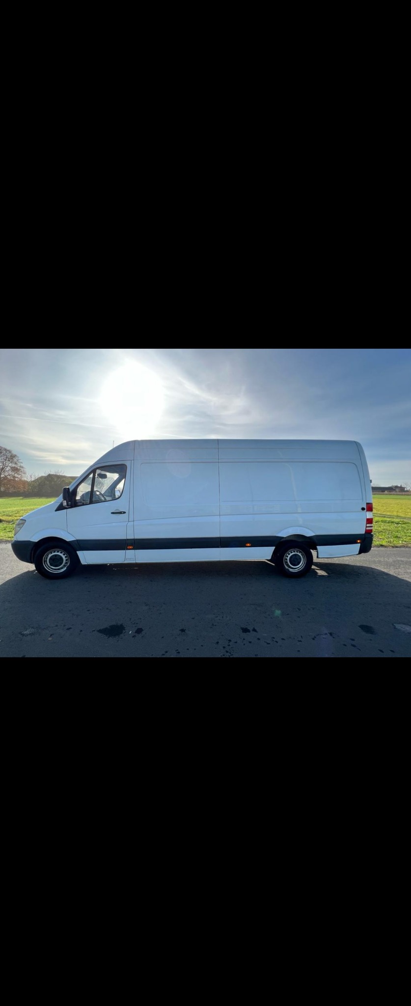 Biały samochód dostawczy Mercedes-Benz Sprinter, widziany z boku na tle słonecznego, błękitnego nieba i zieleni, zaparkowany na asfalcie z widocznymi plamami.