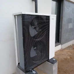 Zewnętrzna jednostka pompy ciepła Hewalex zamontowana na betonowych bloczkach przy ścianie budynku z szarą elewacją i czarną rynną.