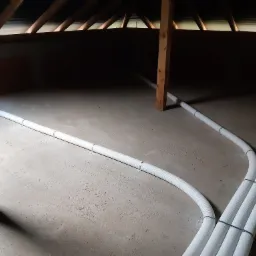 Białe, karbowane rury wentylacyjne prowadzone po betonowej podłodze pod ciemnym stropem z drewnianych belek, mocowane metalowymi obejmami.