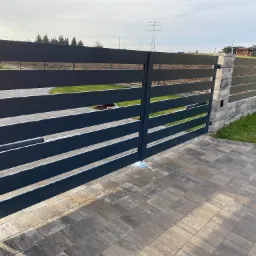 Nowoczesna, pozioma brama wjazdowa z szarych, aluminiowych profili, zamontowana na betonowych słupkach z kostki, widok z zewnątrz na podjeździe z kostki brukowej, w tle trawnik i dom.