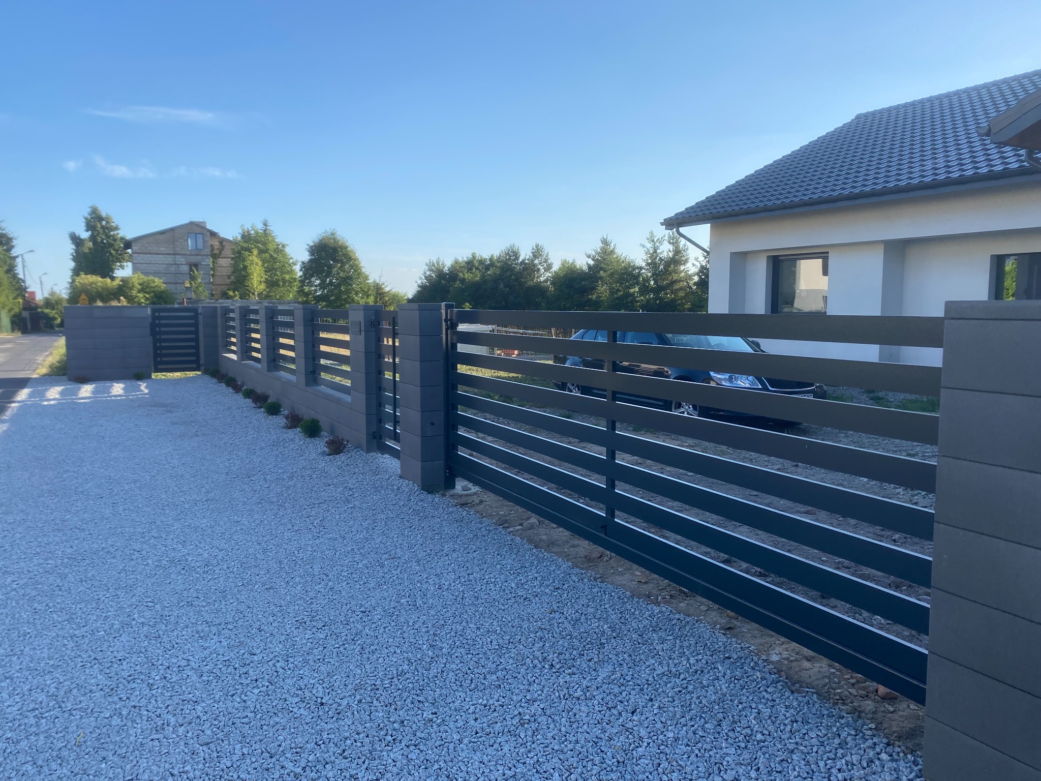 Nowoczesne ogrodzenie panelowe w kolorze grafitowym z przesuwną bramą, widok z zewnątrz na posesję wysypaną białym gresem, w tle dom z ciemną dachówką i zaparkowany samochód.