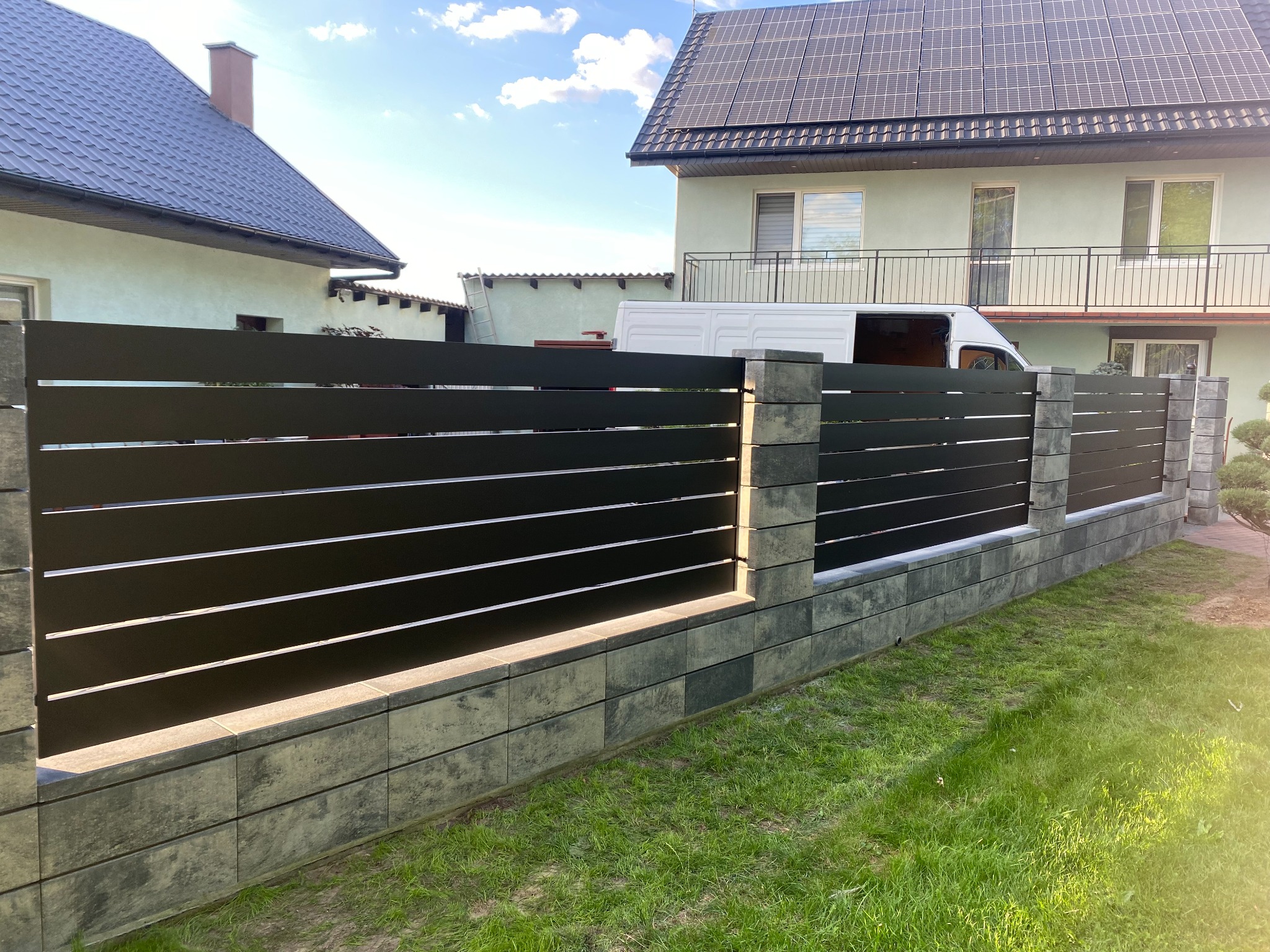 Nowoczesne ogrodzenie panelowe z poziomych, czarnych listew i betonowych słupków w odcieniach szarości, na tle zadbanego trawnika i dwóch domów, z których jeden ma panele słoneczne na dachu, a za...