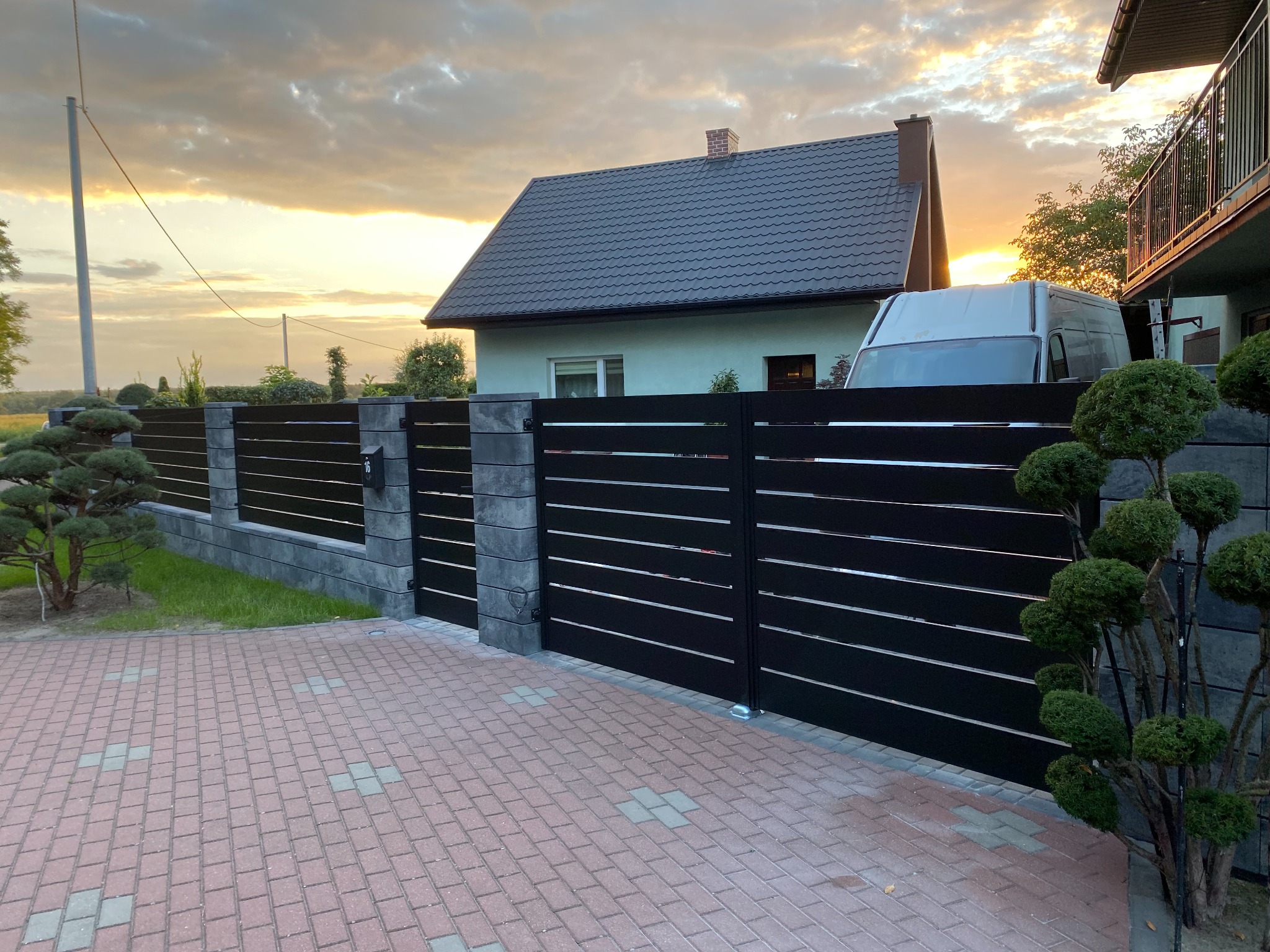 Nowoczesne ogrodzenie panelowe z czarnymi przęsłami i szarymi słupkami betonowymi, brama wjazdowa, kostka brukowa na podjeździe, w tle dom jednorodzinny i zachodzące słońce.