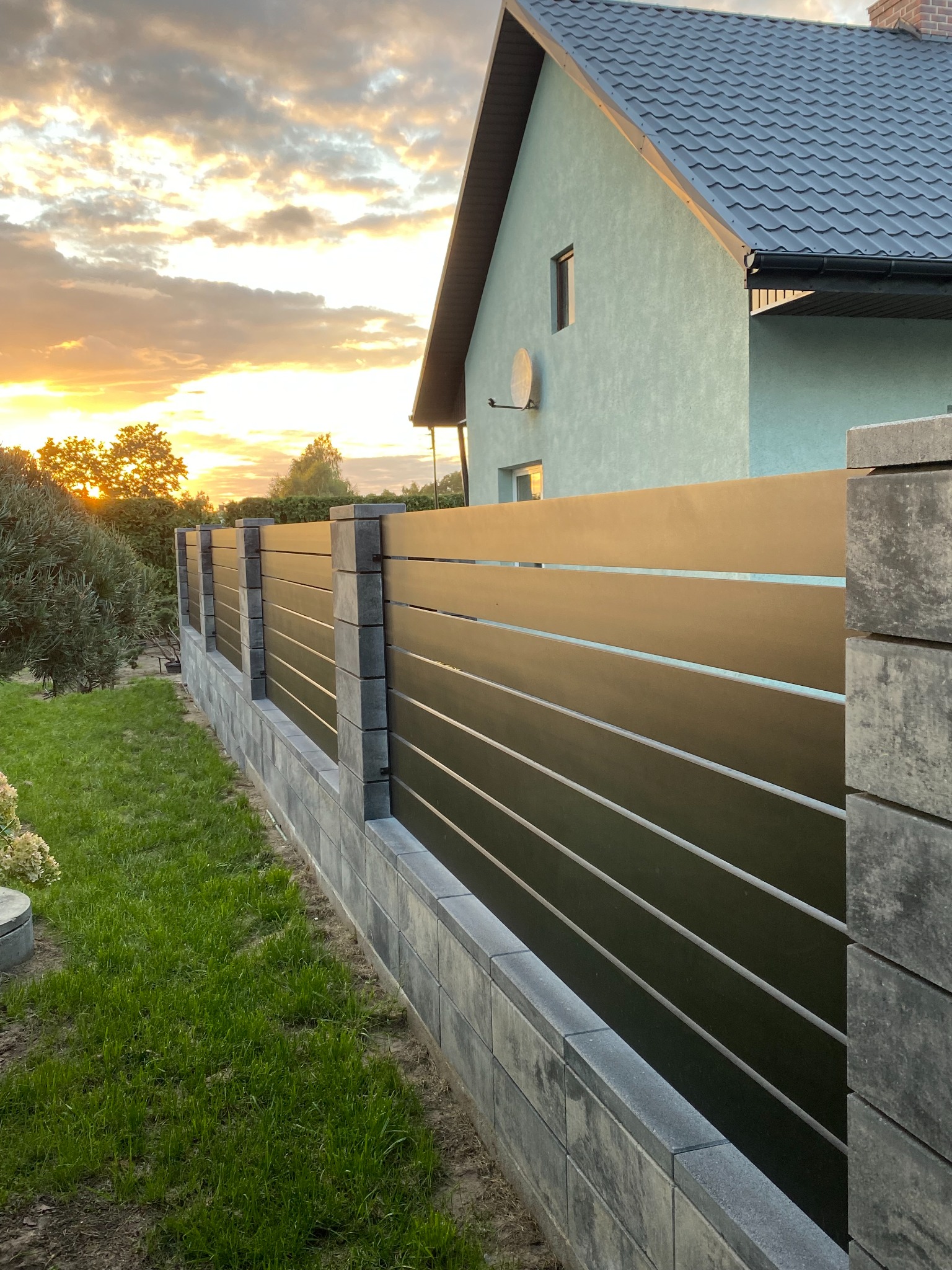 Nowoczesne ogrodzenie panelowe z poziomych, brązowych desek na fundamencie z szarej kostki betonowej, na tle domu z szarą dachówką i zachodzącego słońca.