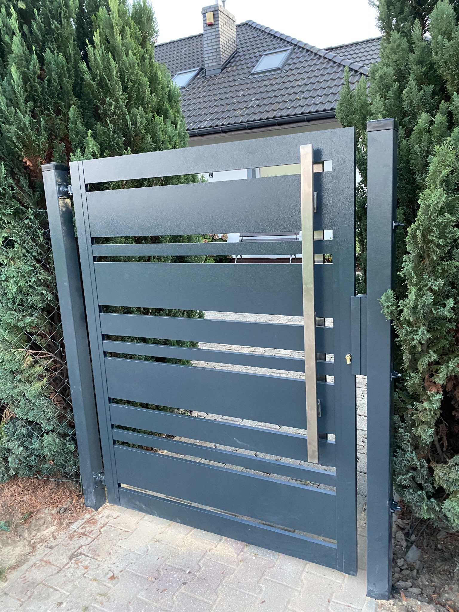 Nowoczesna, antracytowa brama posesyjna z poziomymi panelami, stalowy uchwyt, widok z zewnątrz, kostka brukowa, tuje w tle.