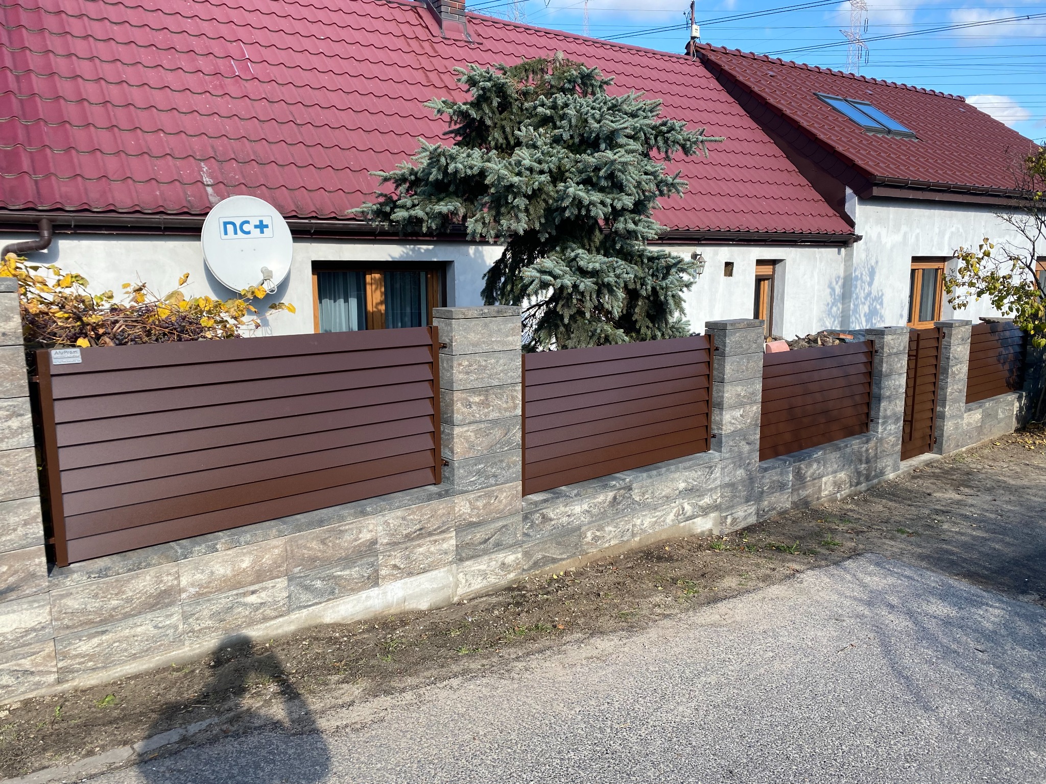 Nowoczesne ogrodzenie panelowe w kolorze brązowym, z podmurówką z szarej kostki, na tle domu z czerwoną dachówką i zielonym świerkiem.