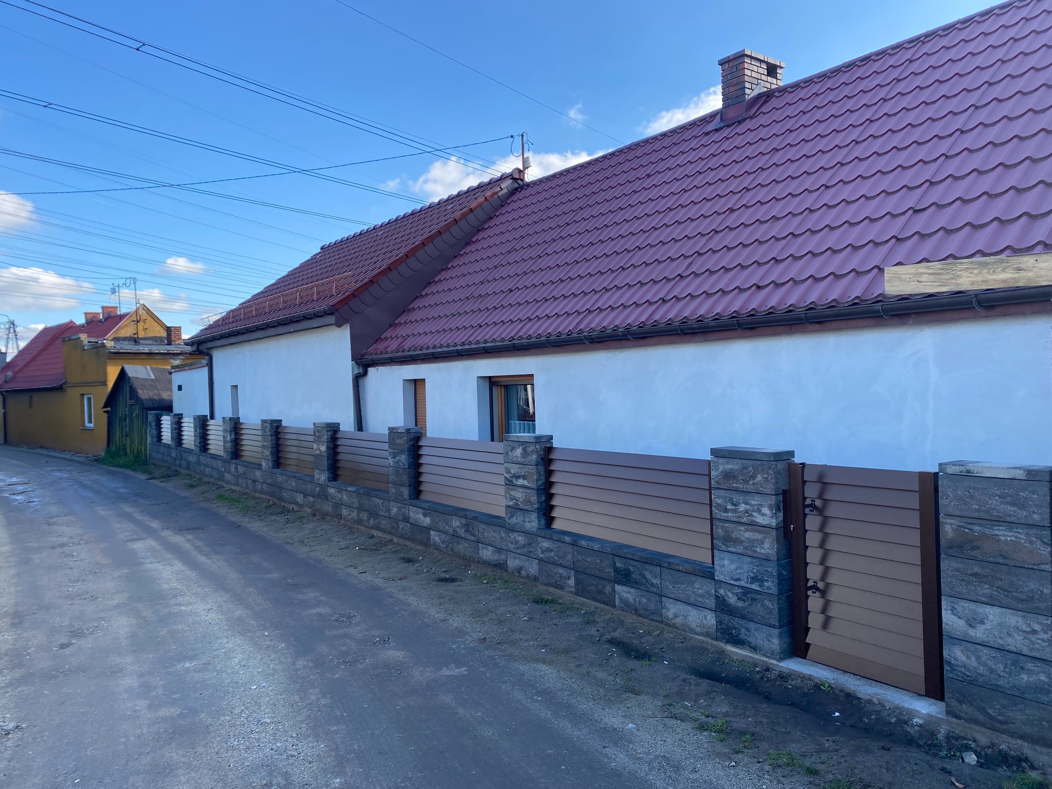 Nowoczesne ogrodzenie panelowe z poziomych desek w kolorze brązowym i kamiennych słupków, brama wejściowa, dom z czerwoną dachówką w tle, widok z zewnątrz.