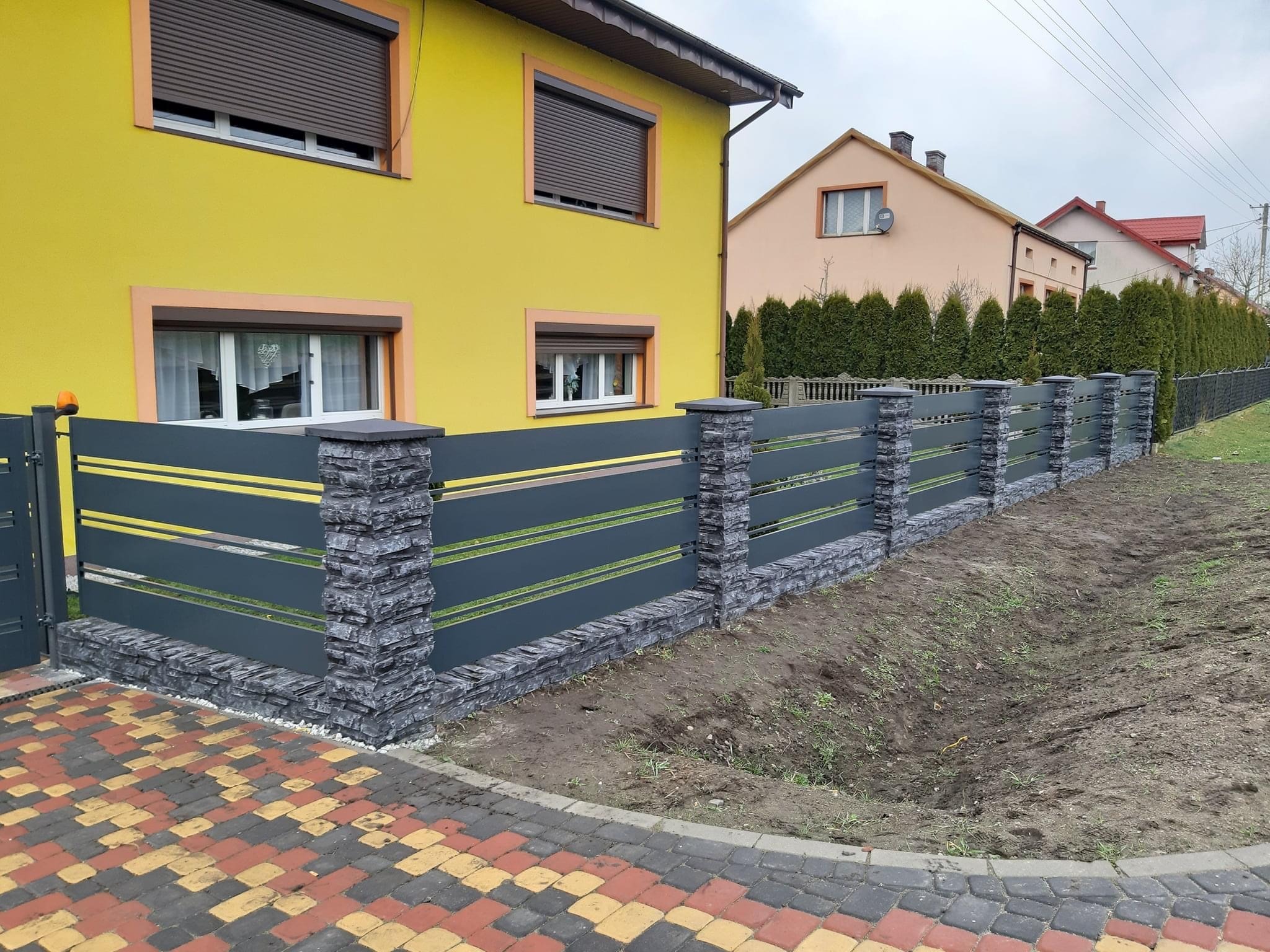 Nowoczesne ogrodzenie panelowe w kolorze grafitowym z elementami imitującymi kamień, zamontowane przy żółtym domu z brązowymi roletami, kostka brukowa na pierwszym planie.
