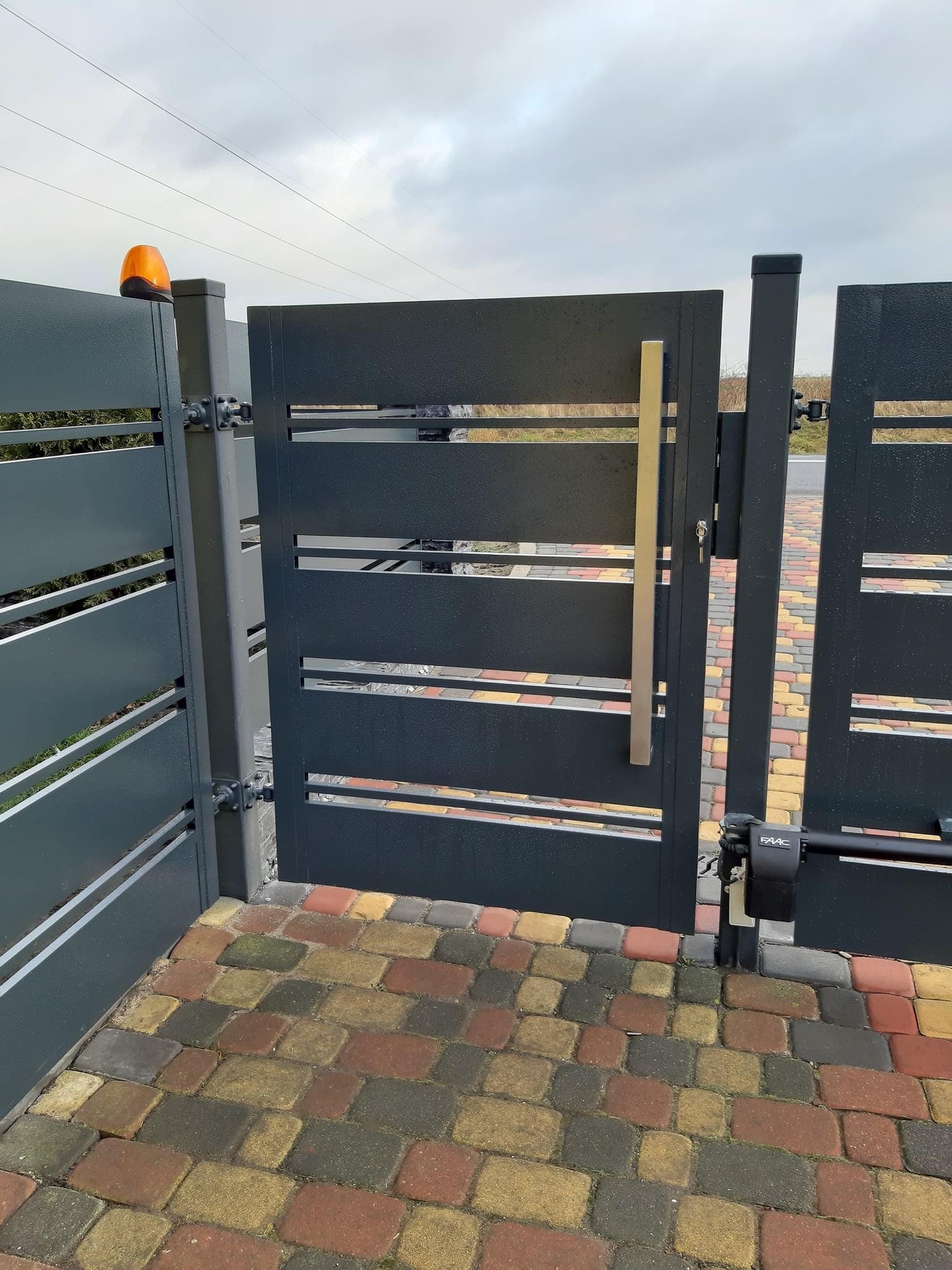 Nowoczesna, grafitowa brama wjazdowa z automatyką i pomarańczową lampą ostrzegawczą, zamontowana w Koninie, widoczna na tle kostki brukowej. Brama częściowo otwarta, z widocznym mechanizmem napędowym.