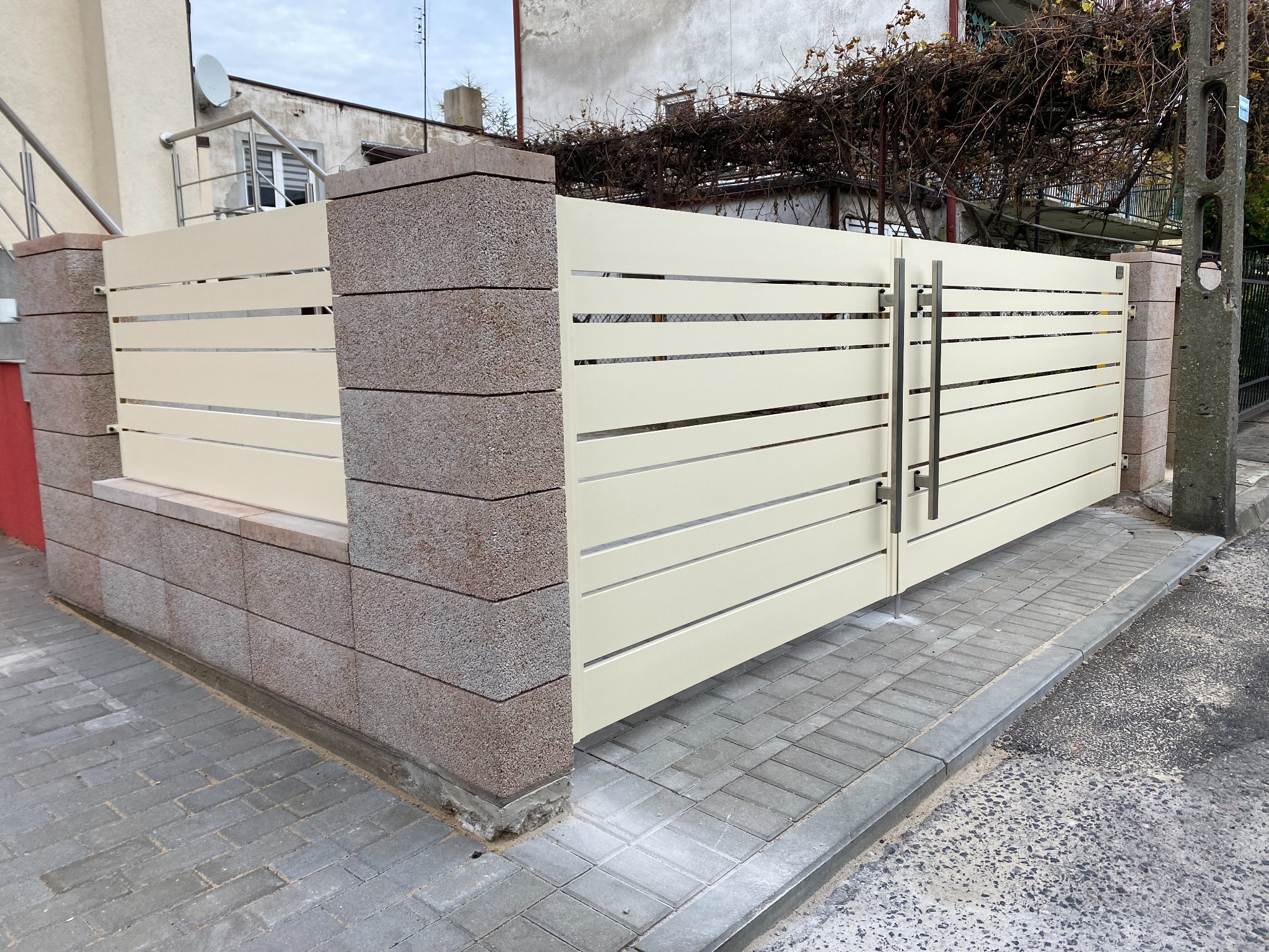 Nowoczesna brama wjazdowa z poziomych profili w kolorze kremowym, osadzona na murowanych słupkach z bloków imitujących kamień, widok z zewnątrz.