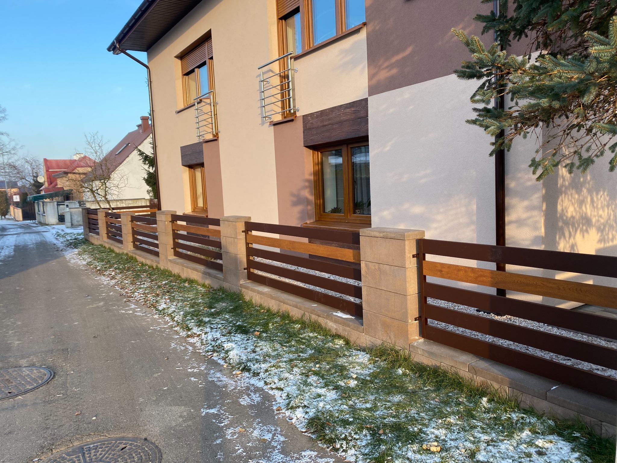Nowoczesne ogrodzenie panelowe z brązowych desek i kamiennych słupków, przylegające do elewacji domu o beżowo-brązowej kolorystyce, z resztkami śniegu na trawie i chodniku.