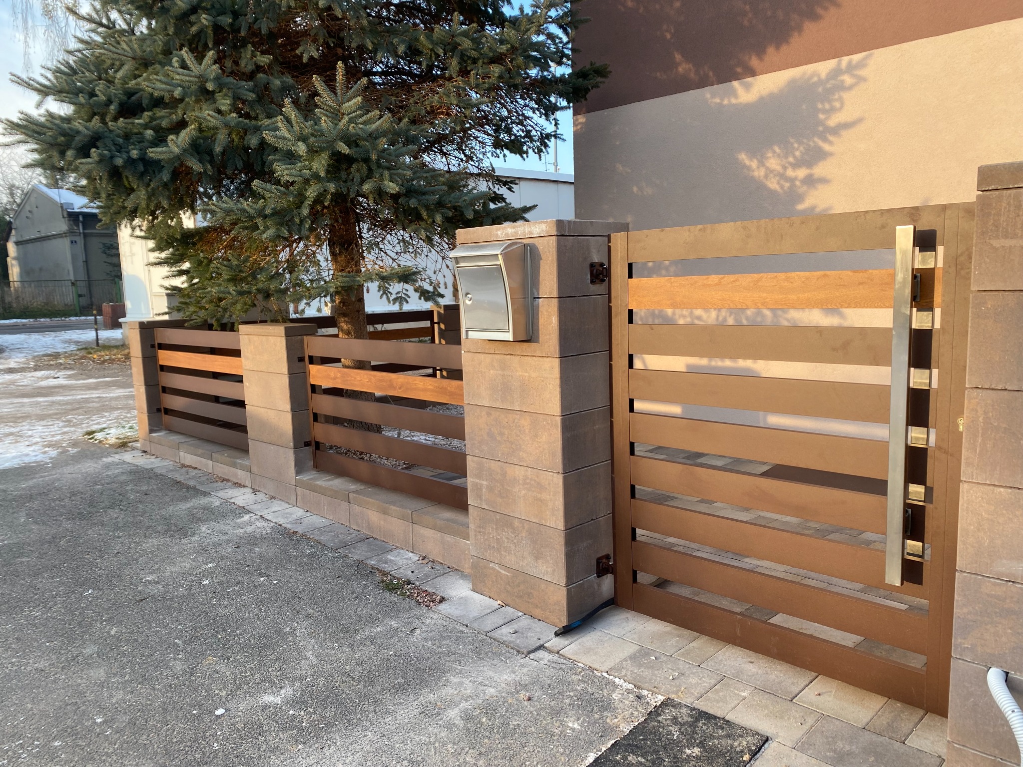 Nowoczesne ogrodzenie panelowe z brązowymi przęsłami i słupkami z szarej kostki, brama wjazdowa z pionowym, metalowym uchwytem, srebrna skrzynka na listy zamontowana na słupku, w tle iglaste drzewo...