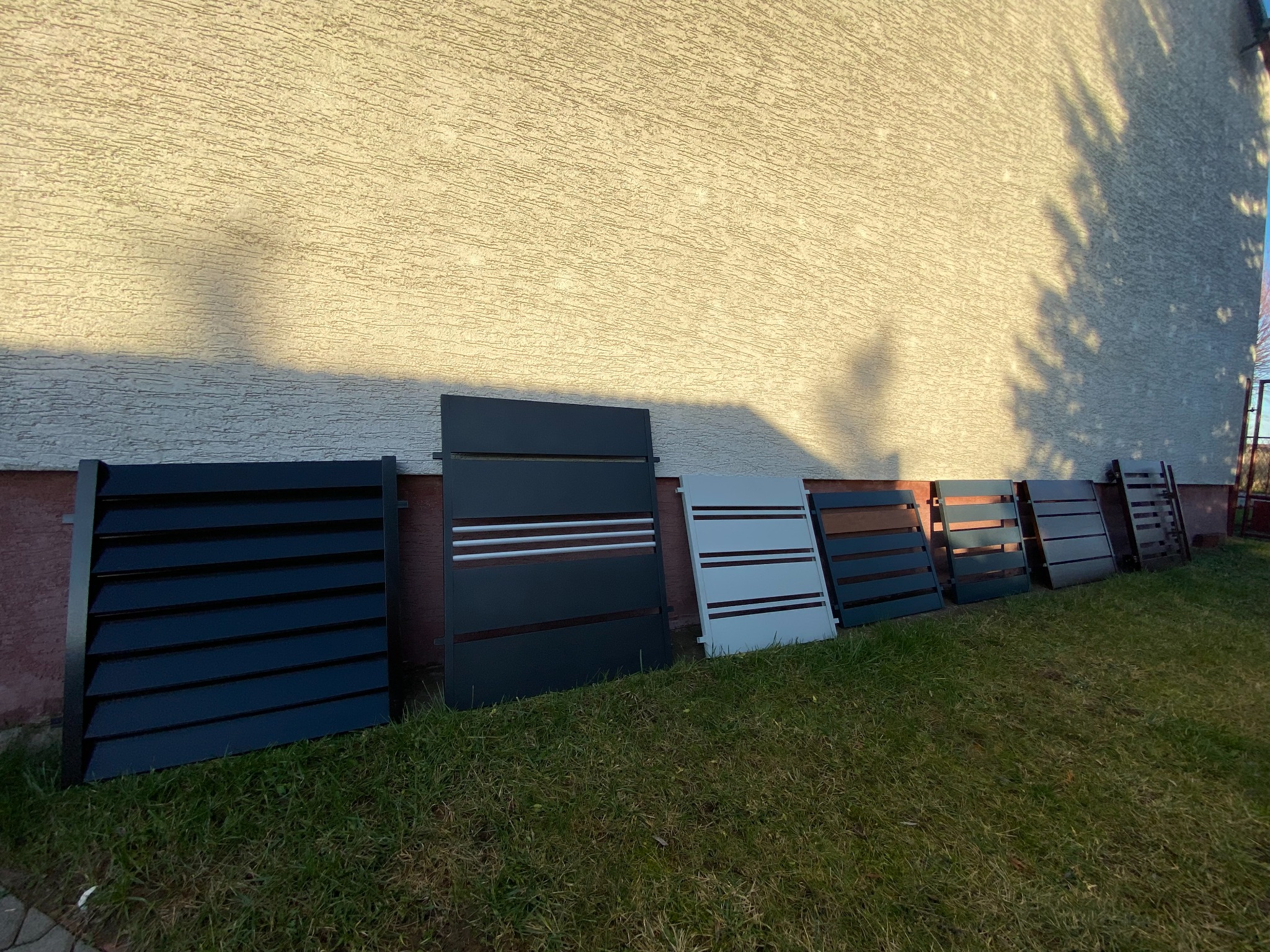 Panele ogrodzeniowe w różnych kolorach i wzorach, oparte o ścianę budynku z fakturą, na trawniku w słoneczny dzień.