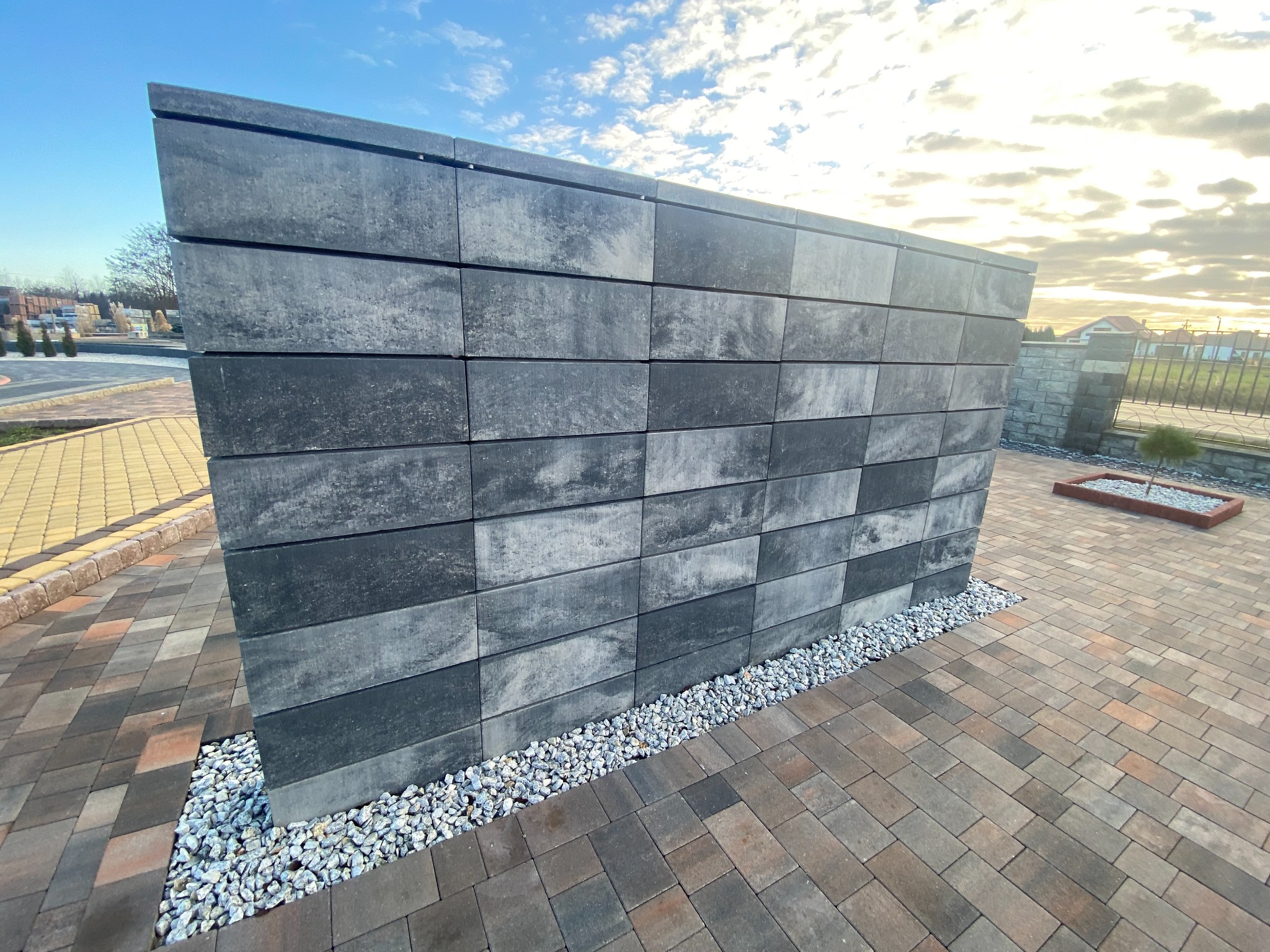 Nowoczesny mur z szarej kostki betonowej ułożonej poziomo, oddzielony od kostki brukowej białym grysem, na tle błękitnego nieba z chmurami.