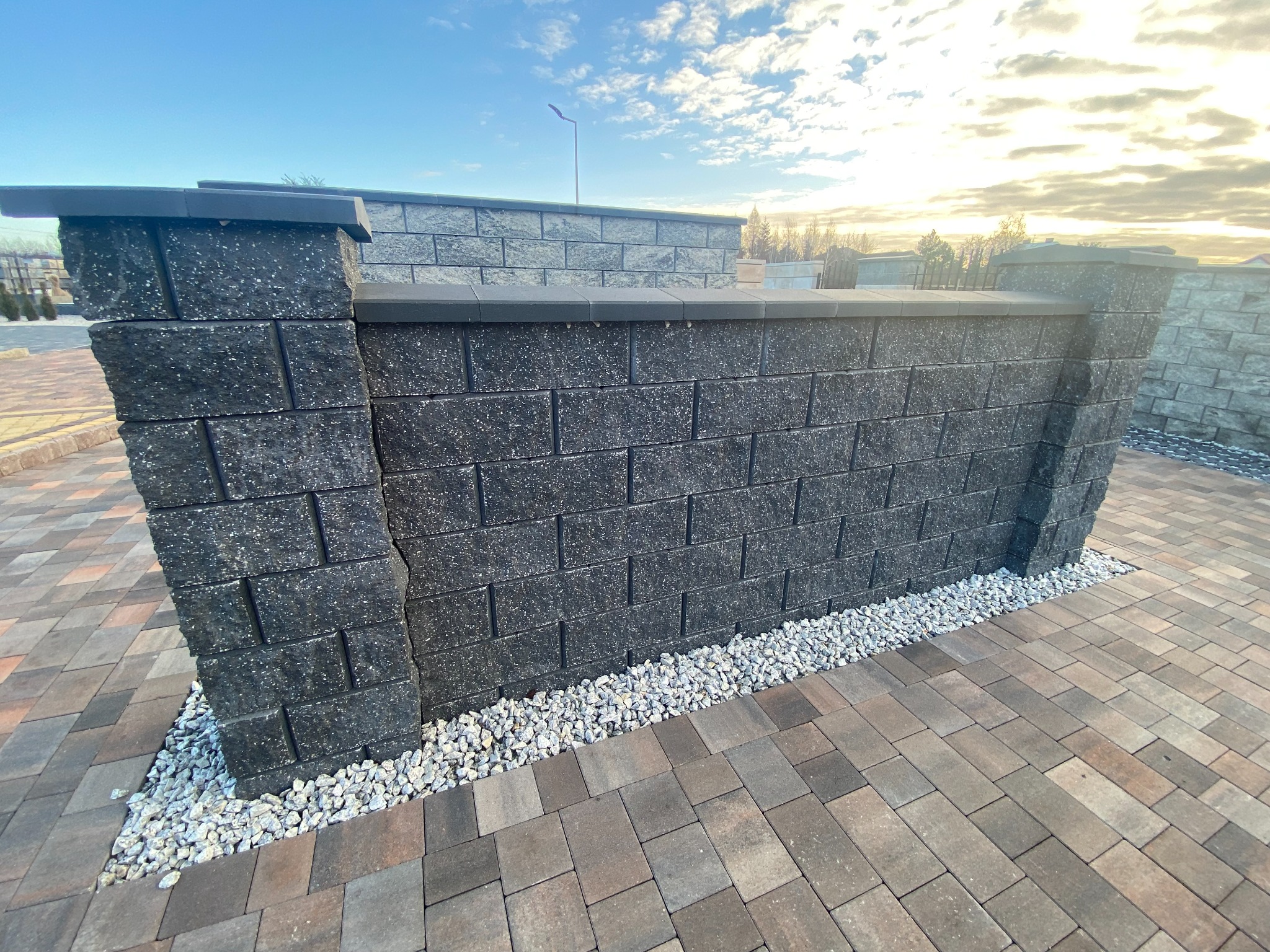 Nowoczesny, niski murek z ciemnoszarej kostki łupanej, wykończony poziomą listwą i kapą, oddzielający przestrzeń wyłożoną kostką brukową, z podsypką z białego grysu, w tle widok na zachodzące słońce.