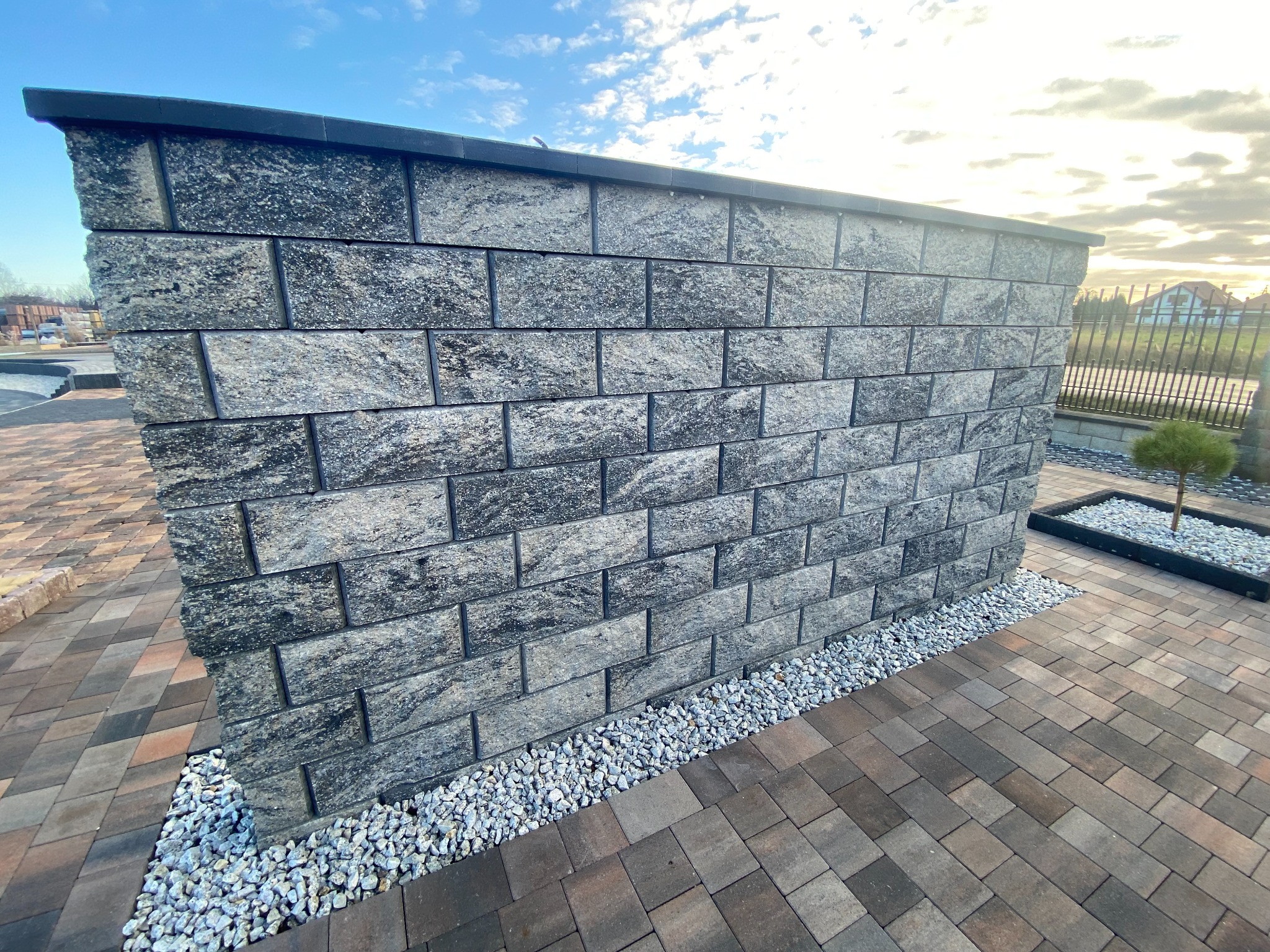 Mur oporowy z szarej kostki łupanej, wykończony drobnym białym żwirem, na tle kostki brukowej i fragmentu ogrodzenia.