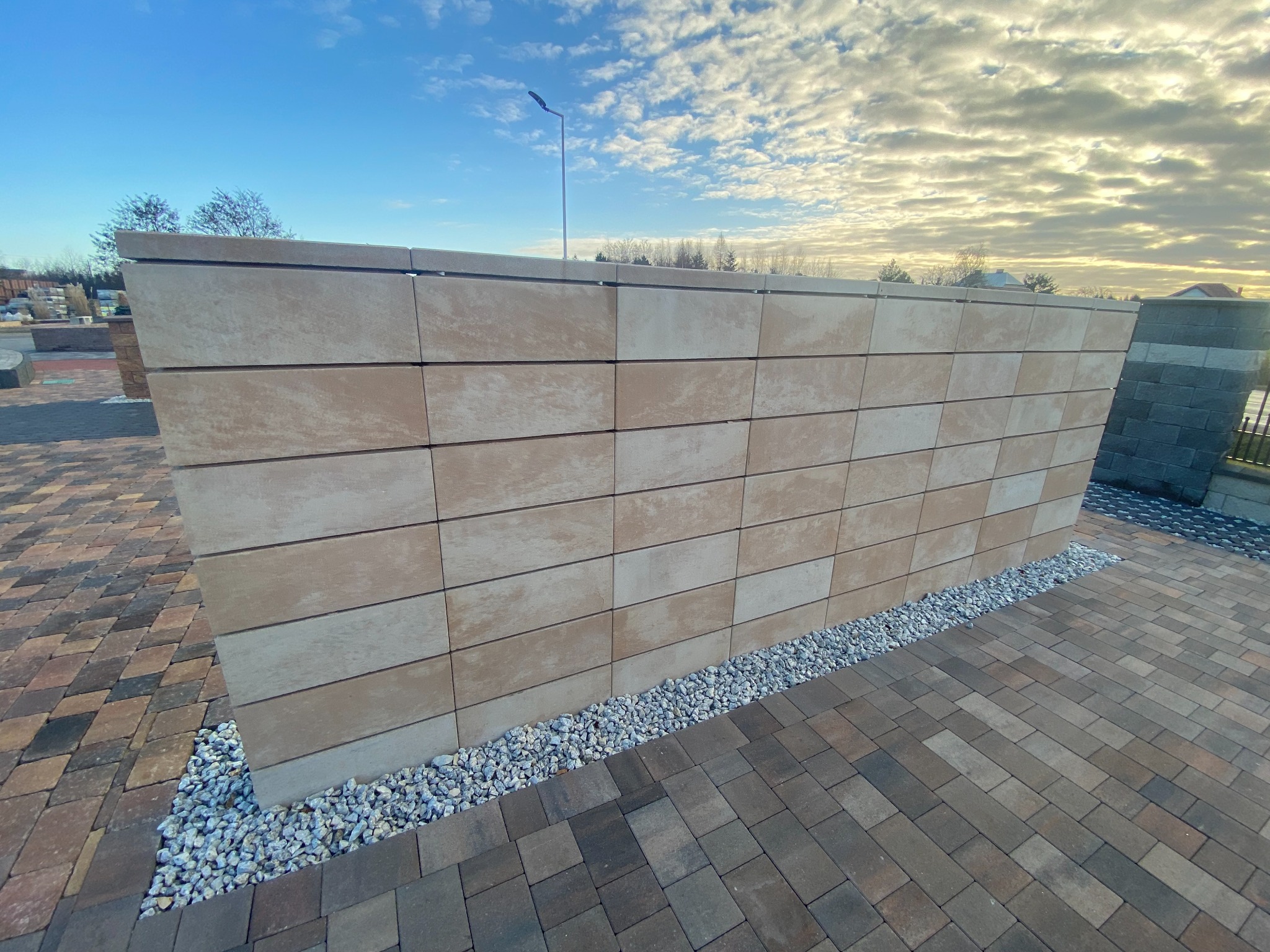 Nowoczesny, beżowy murek ogrodzeniowy z prefabrykatów betonowych, oddzielony od kostki brukowej pasem białego grysu, na tle błękitnego nieba z chmurami.