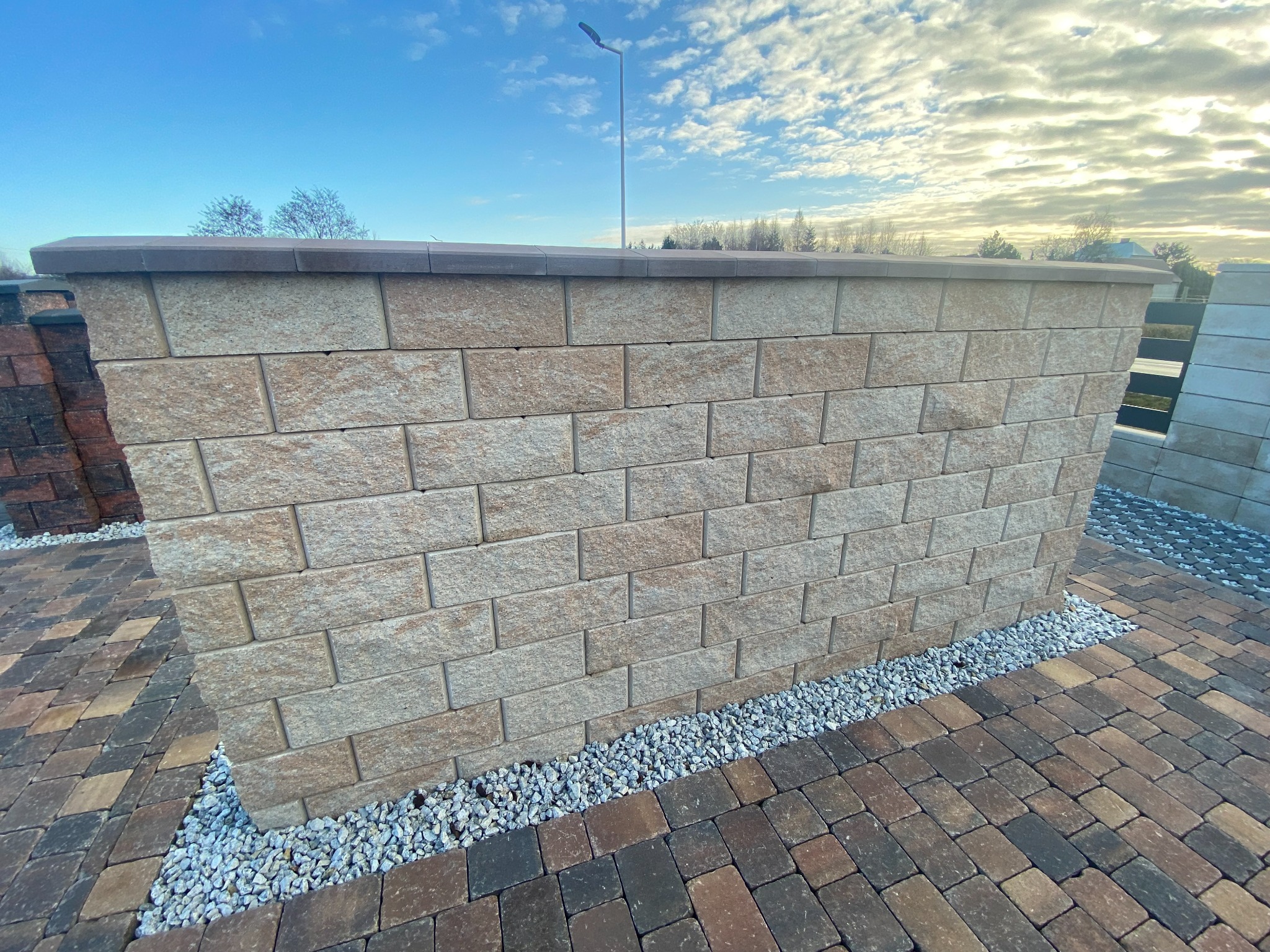 Fragment murku ogrodzeniowego z bloczków betonowych o piaskowej barwie, wykończonego ciemniejszą nakrywą, ułożony na kostce brukowej z jasnymi kamykami u podstawy, na tle błękitnego nieba z chmurami.