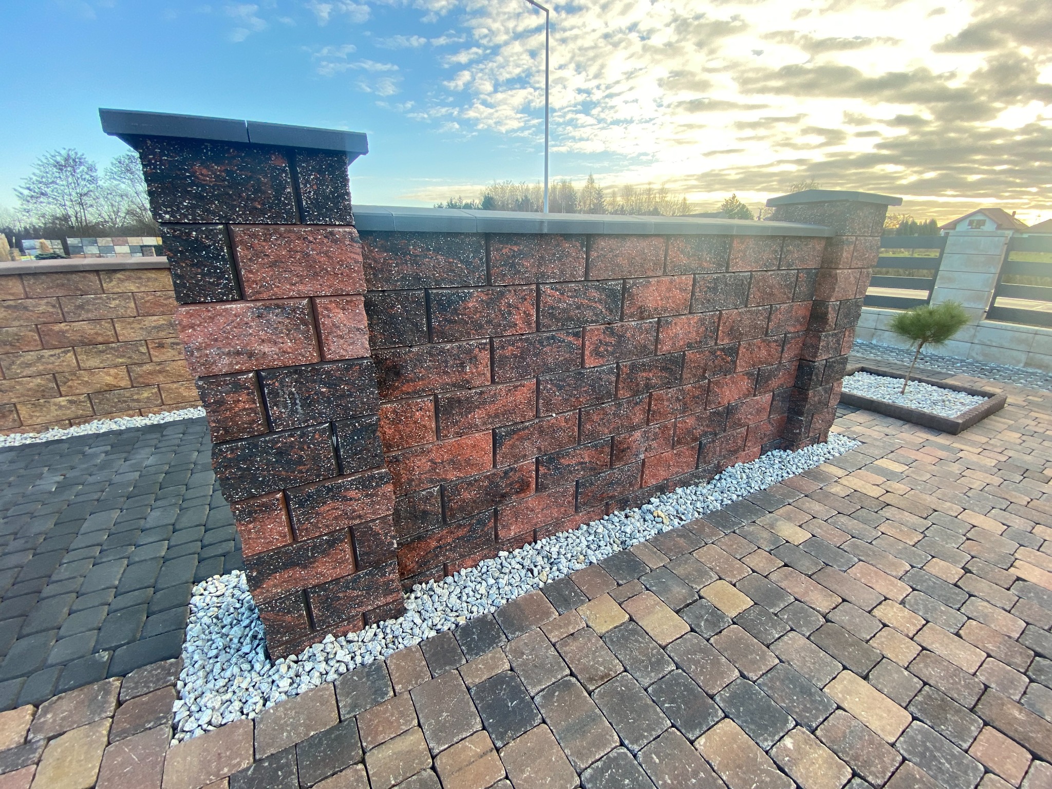 Fragment murku ogrodzeniowego z czerwono-brązowych bloczków z czarnymi akcentami, zwieńczonego szarym daszkiem, z drobnym białym grysem u podstawy, na tle kostki brukowej w odcieniach brązu...