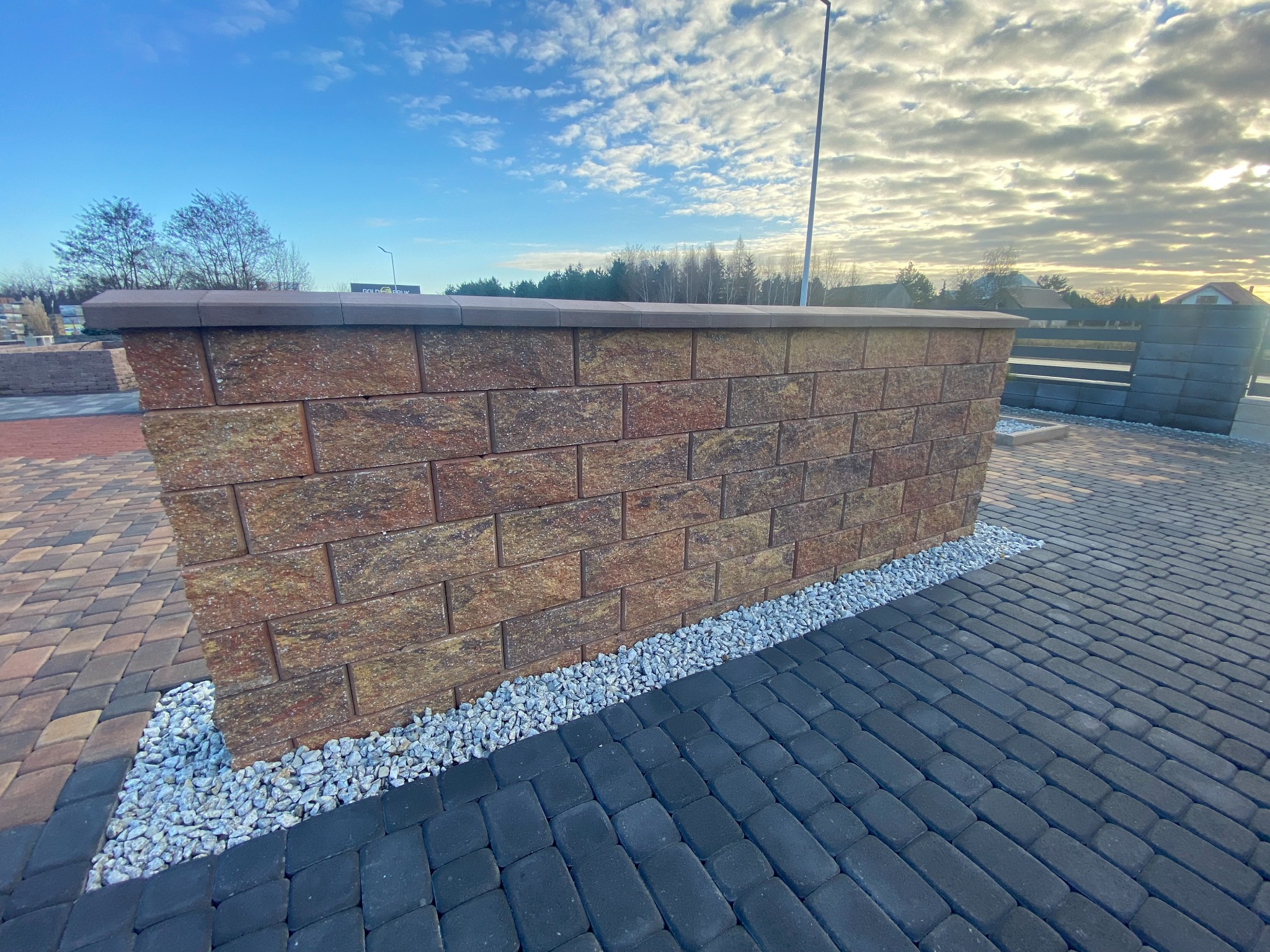 Fragment ogrodzenia z cegieł klinkierowych z brązowym daszkiem, ozdobiony białym grysem, na tle kostki brukowej w odcieniach szarości i brązu, oświetlony popołudniowym słońcem.